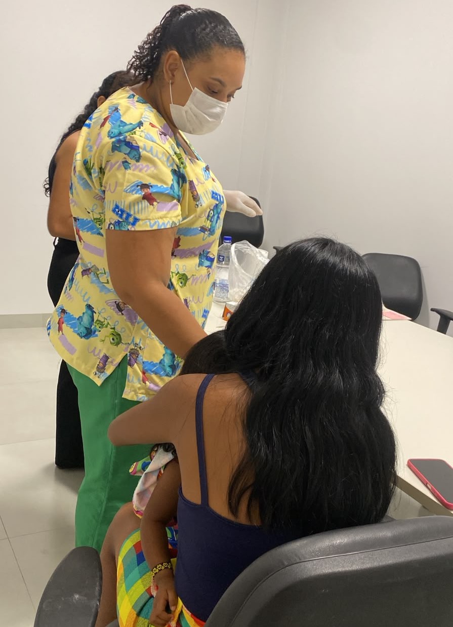  A imagem mostra uma profissional de saúde realizando atendimento a uma mulher com uma criança no colo, durante uma coleta de material genético em uma sala de atendimento.  A profissional está em pé, usa máscara, luvas, calça verde e jaleco estampado em tons de amarelo e azul. À sua frente, a mulher está sentada de costas para a câmera, com cabelos longos e pretos, vestindo uma blusa azul de alças, enquanto segura uma criança pequena no colo, parcialmente coberta por um pano colorido. Ao fundo, há outra pessoa parcialmente visível, além de uma mesa com frascos e materiais de atendimento, cadeiras pretas e paredes brancas, compondo um ambiente clínico e organizado.