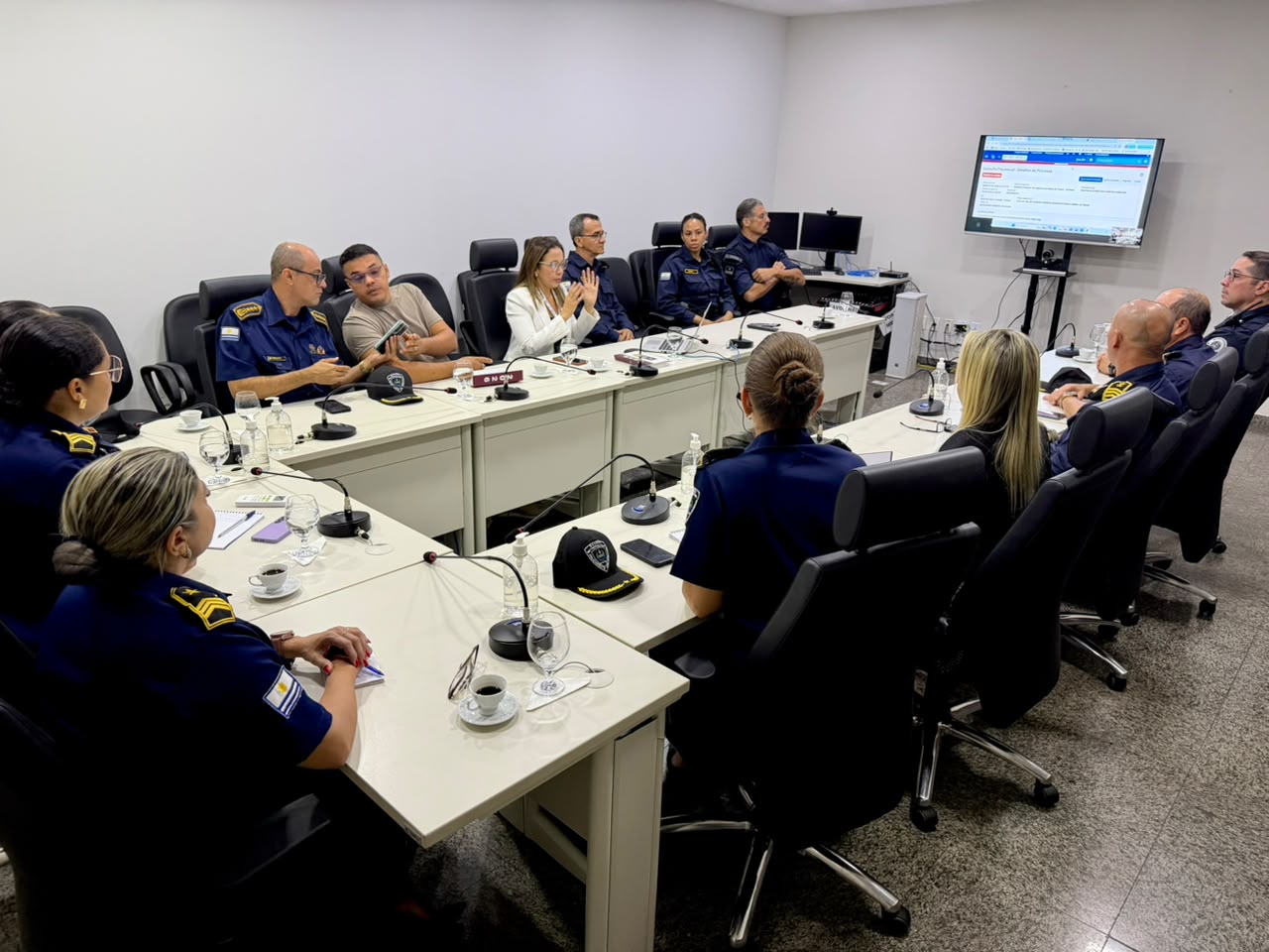 Reunião em ambiente institucional, com diversos participantes sentados ao redor de uma mesa em U, incluindo integrantes da área de segurança