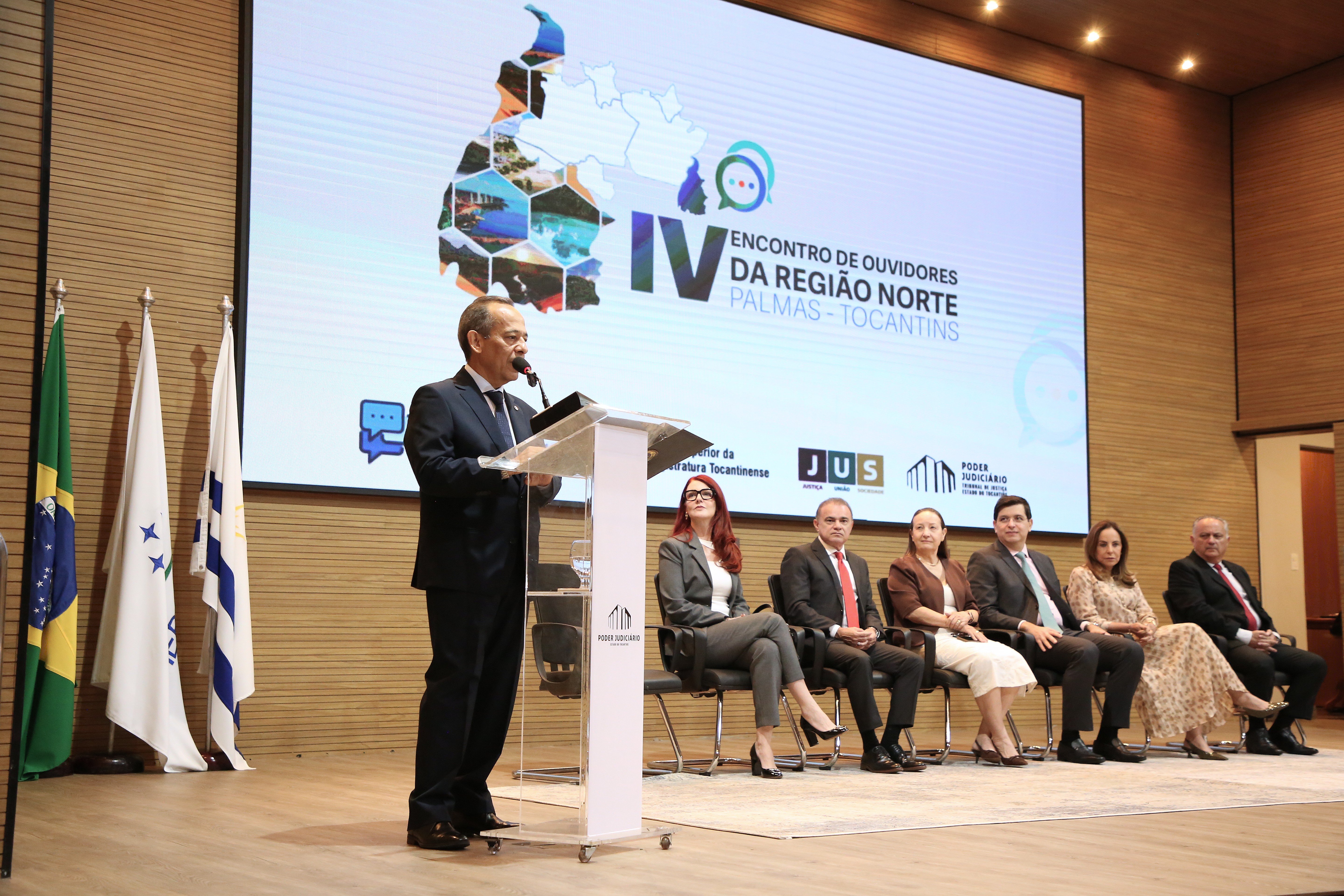 O desembargador João Rodrigues discursa no púlpito durante o IV Encontro de Ouvidores e Ouvidoras Judiciais da Região Norte. Ao fundo, autoridades acompanham a solenidade no palco e o painel oficial do evento.