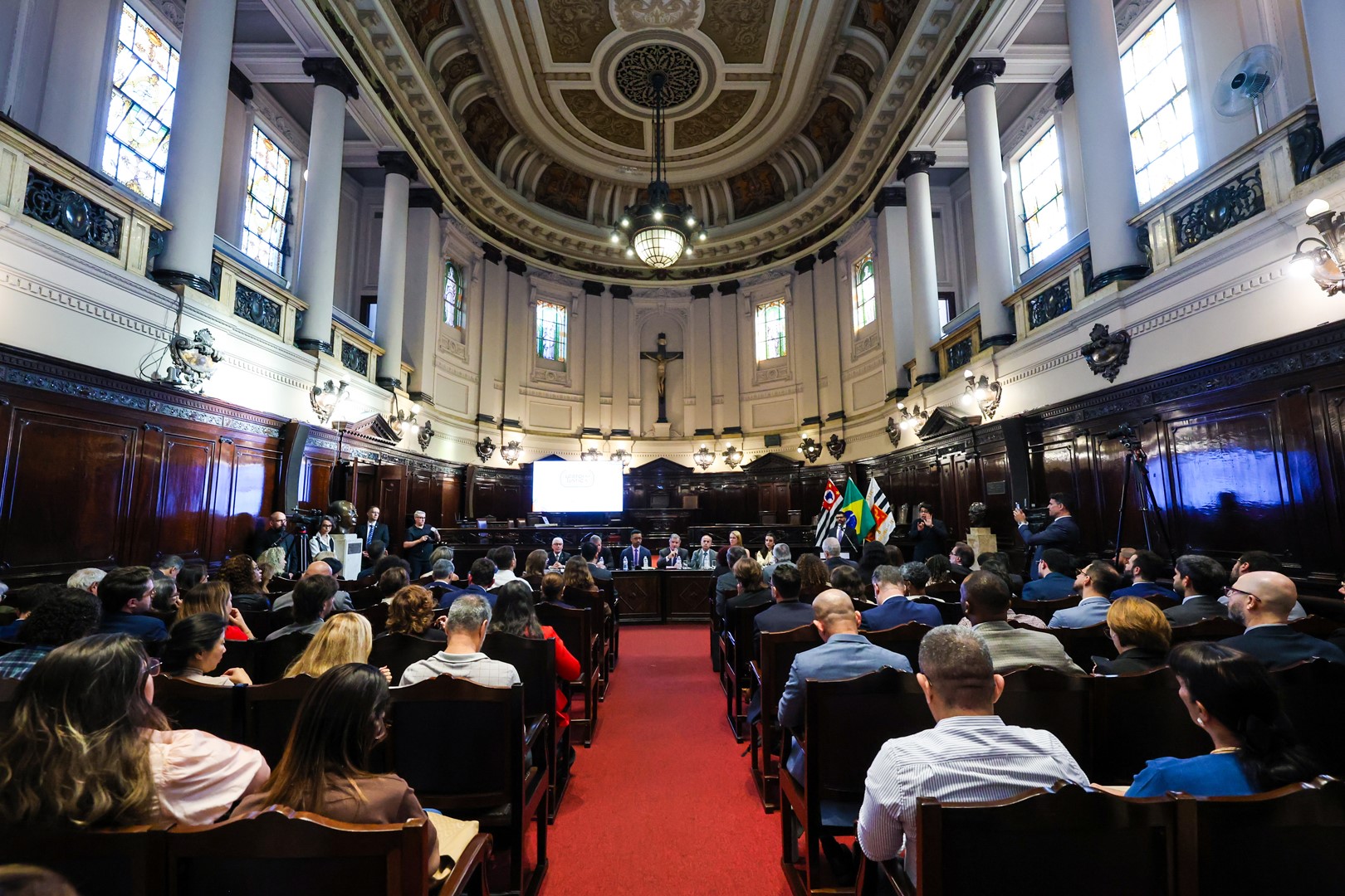 Auditório com teto ornamentado, vitrais, lustres e colunas, reúne autoridades e participantes do 2º Encontro Nacional do CNJ de Promoção dos Direitos LGBTQIA+ e Acesso à Justiça. Ao centro, integrantes compõem a mesa de abertura; ao fundo, aparecem as bandeiras do Estado de São Paulo, do Brasil e do Tribunal de Justiça de São Paulo.