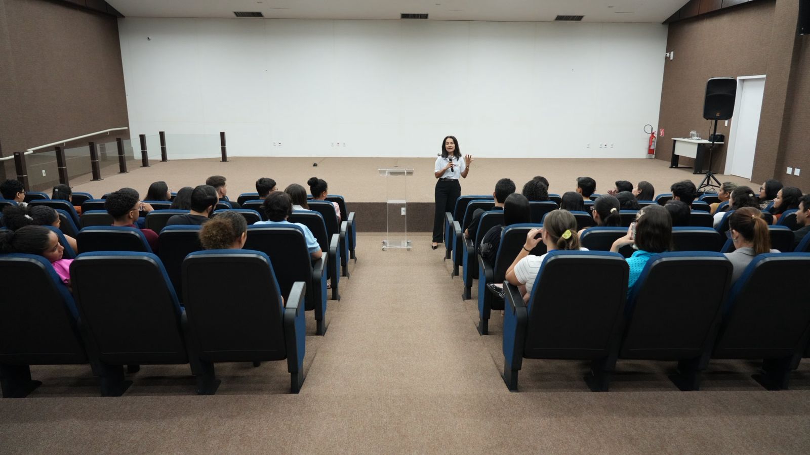 A desembargadora Hélvia Túlia fala ao microfone e gesticula diante do público durante a roda de conversa do projeto “Cassédio Itinerante”, no auditório do Fórum de Araguaína. Os participantes acompanham a atividade sentados em poltronas dispostas em frente ao palco.