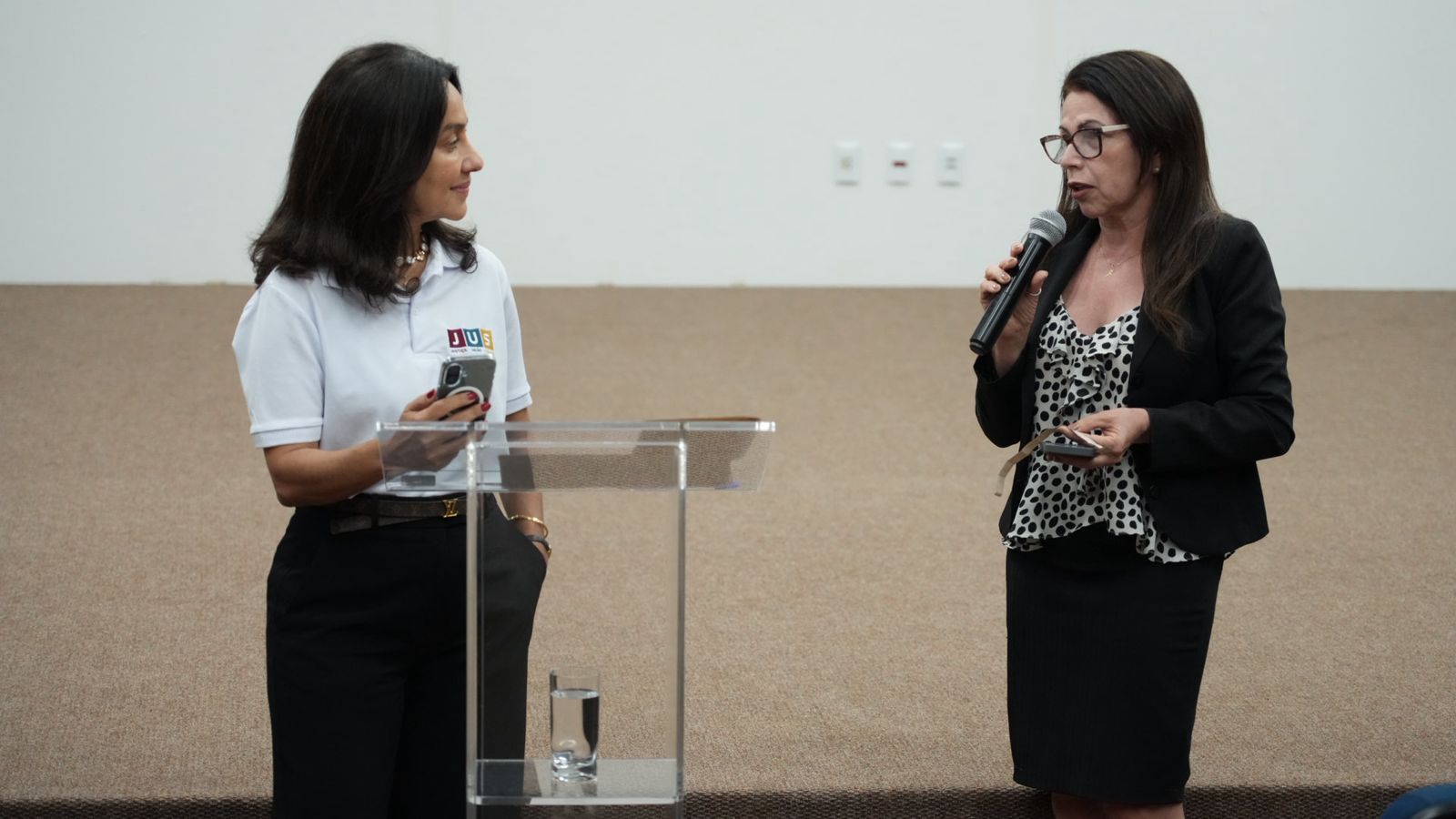 A desembargadora Hélvia Túlia, à esquerda, observa a servidora Márcia, que fala ao microfone durante a roda de conversa do projeto “Cassédio Itinerante”, no Fórum de Araguaína. Ao centro, há um púlpito de acrílico.