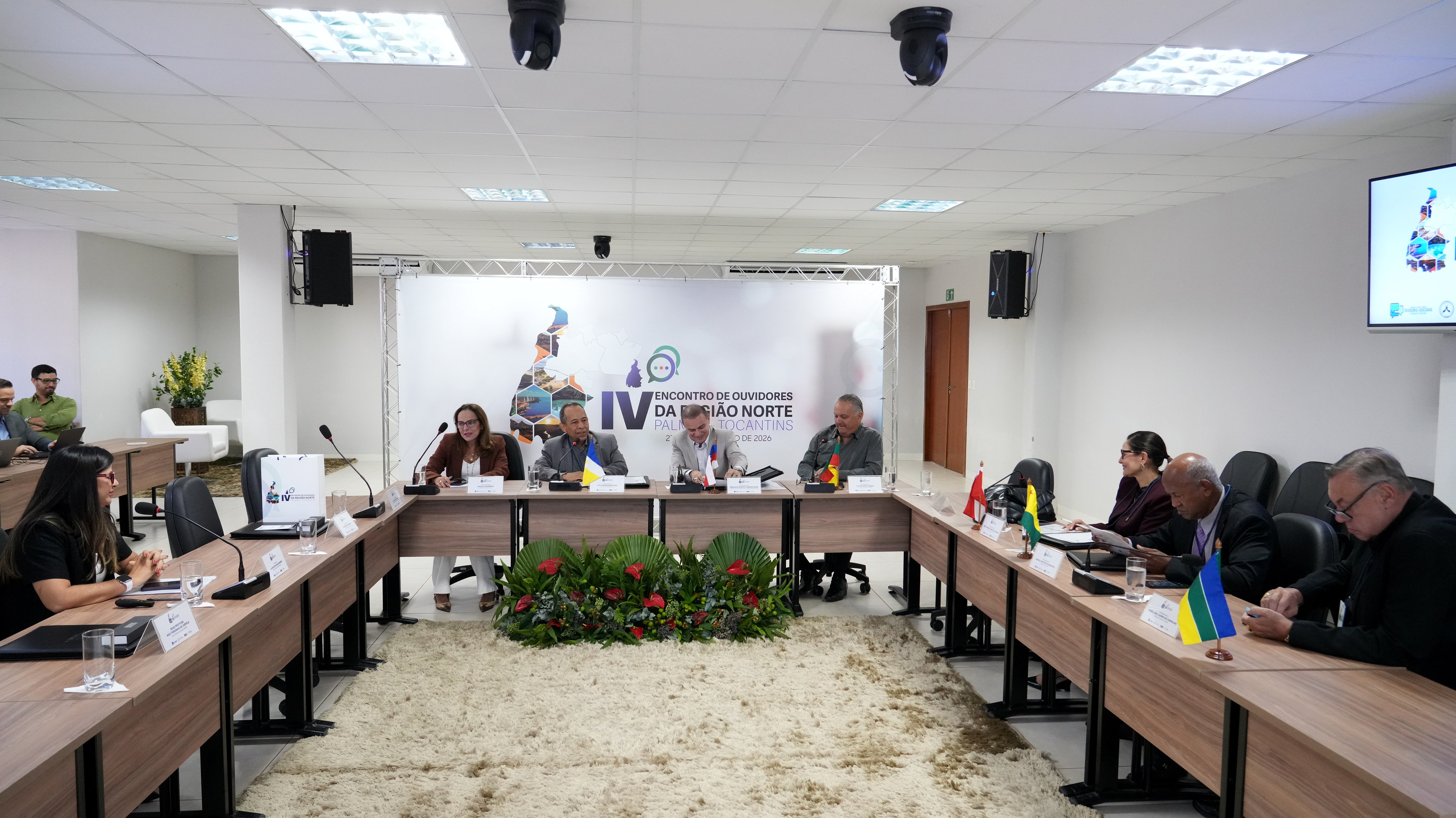A imagem mostra uma reunião realizada na sala da Escola Superior da Magistratura Tocantinense, ESMAT, durante o IV Encontro de Ouvidores da Região Norte, em Palmas. As mesas estão dispostas em formato de U, com oito participantes sentados, entre homens e mulheres, usando trajes sociais. Ao centro, quatro representantes compõem a mesa principal, com microfones, placas de identificação e pequenas bandeiras de estados da Região Norte sobre as mesas. Ao fundo, um painel exibe a identidade visual do evento com o mapa da Região Norte e o nome do encontro. 