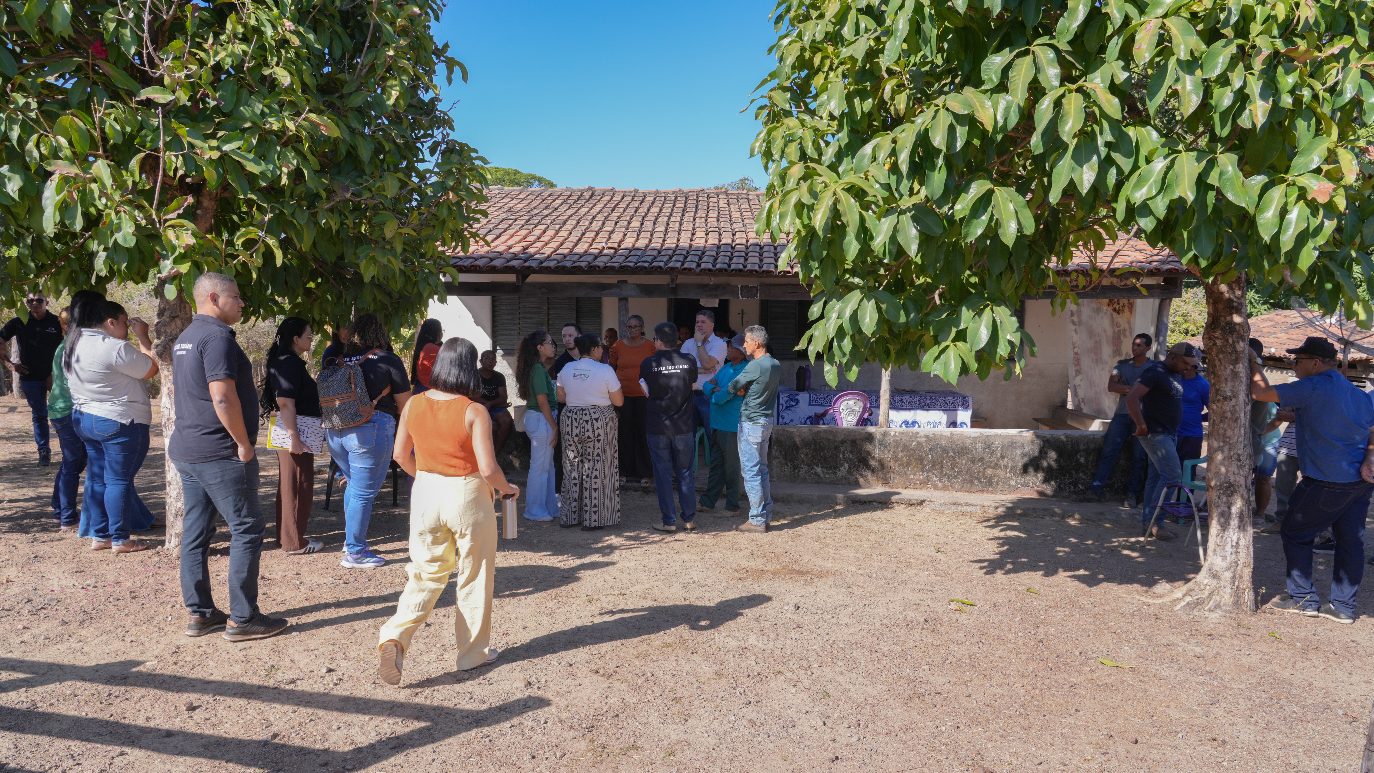 Grupo formado por moradores e representantes de instituições reúne-se em frente a uma casa na área rural durante visita técnica da Comissão Regional de Soluções Fundiárias. As pessoas estão distribuídas sob a sombra de árvores, próximas à varanda da residência, em momento de diálogo e escuta sobre o conflito fundiário.
