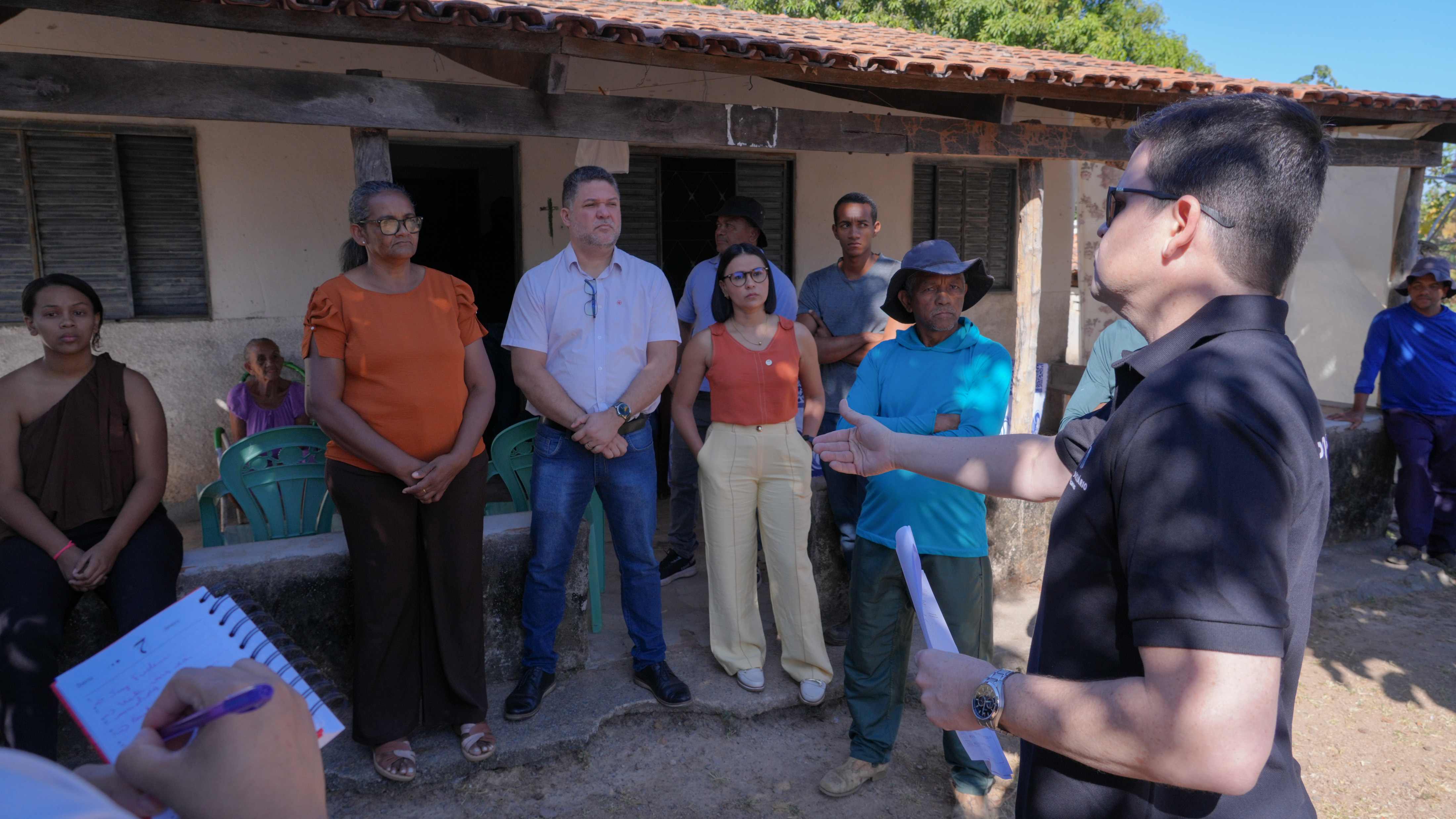 Juiz Frederico Paiva Bandeira de Souza conversa com moradores e representantes de instituições durante visita técnica da Comissão Regional de Soluções Fundiárias. O grupo está reunido em frente a uma residência na área rural. O magistrado segura documentos e gesticula enquanto fala aos participantes.