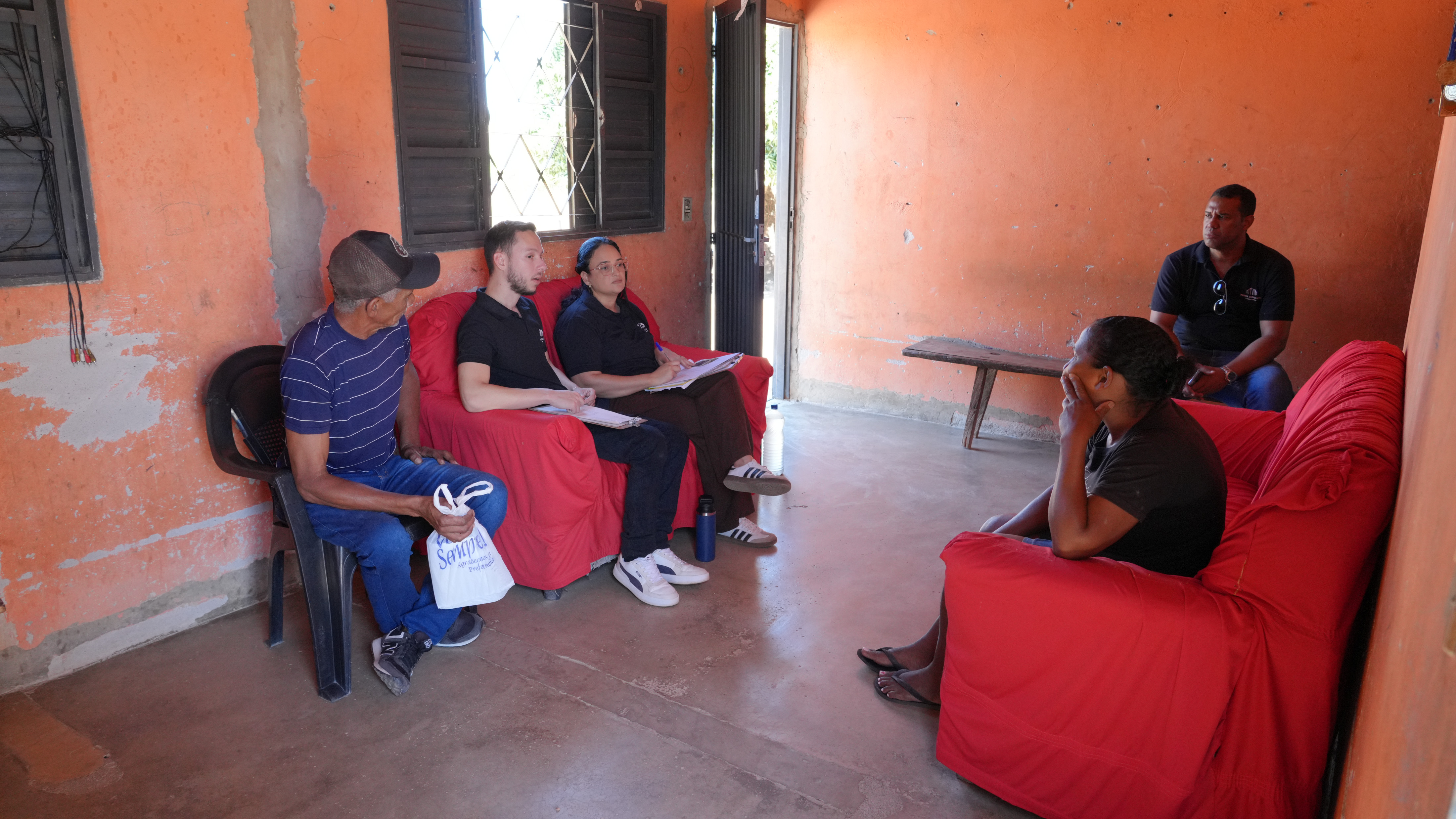 Em uma varanda de paredes alaranjadas, integrantes da equipe técnica conversam com moradores durante visita a uma das áreas em conflito fundiário. Cinco pessoas aparecem sentadas em cadeiras e sofás, enquanto integrante da equipe fazem anotações. A cena registra o momento de escuta e levantamento de informações junto às famílias.