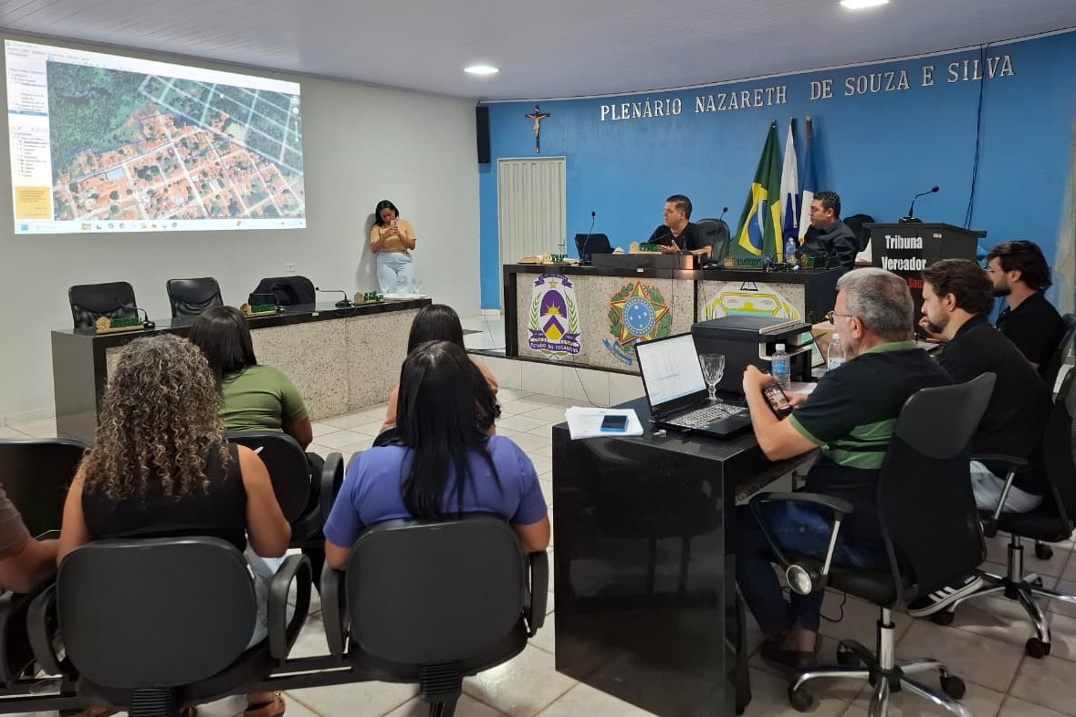 Reunião em plenário, com participantes sentados de frente para uma mesa de autoridades. Um mapa aéreo é projetado em uma tela, enquanto integrantes da mesa acompanham a apresentação. Ao fundo, estão as bandeiras do Brasil e do Tocantins