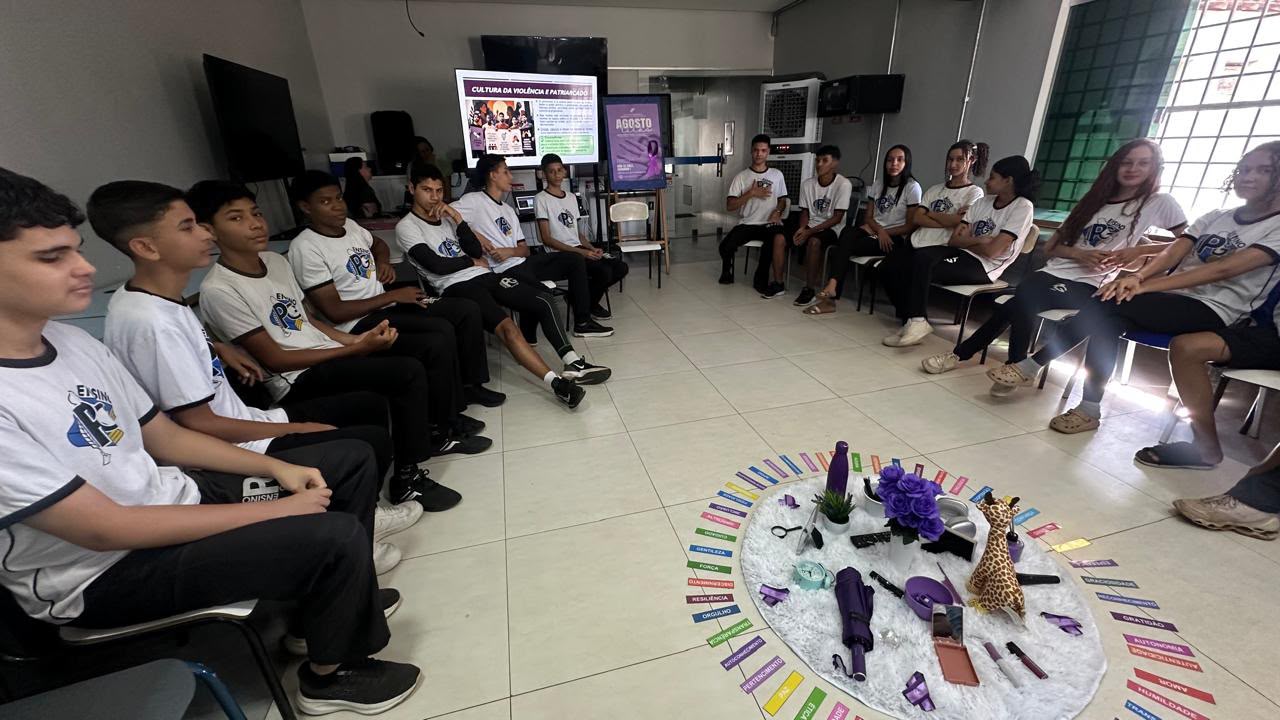 Na sala de aula, estudantes uniformizados estão sentados em círculo durante atividade educativa. Ao centro, objetos e cartões coloridos com palavras compõem uma dinâmica. Ao fundo, uma televisão exibe conteúdo sobre a cultura da violência e o patriarcado.