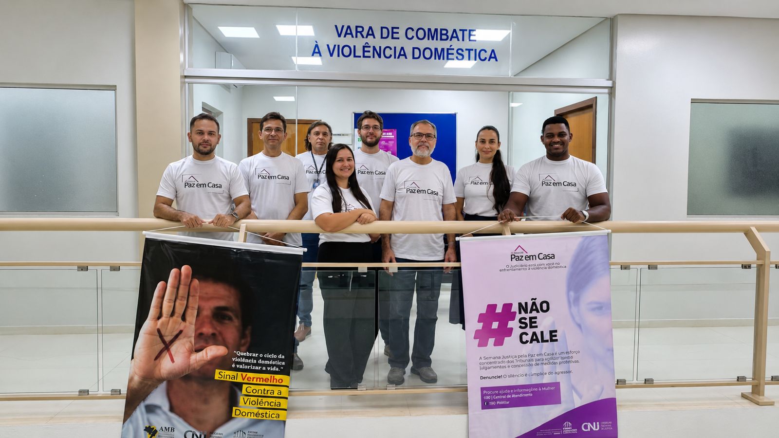 Oito integrantes da equipe posam para foto em frente à Vara de Combate à Violência Doméstica de Palmas. Todos vestem camisetas brancas da campanha Justiça pela Paz em Casa. À frente do grupo, dois banners apresentam mensagens de conscientização e enfrentamento à violência doméstica contra a mulher.
