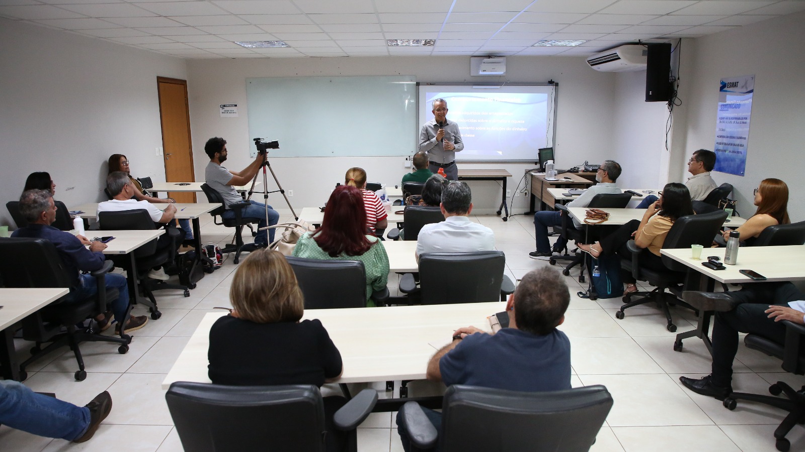 Fotografia da oficina de Preparação para Aposentadoria realizada em uma sala de aula da Esmat, em junho de 2026. Ao fundo, um homem  de camisa cinza fala ao microfone em frente a uma tela de projeção. Os participantes estão sentados em bancadas individuais voltadas para a frente da sala. À esquerda, um fotógrafo com câmera em um tripé registra o evento. 