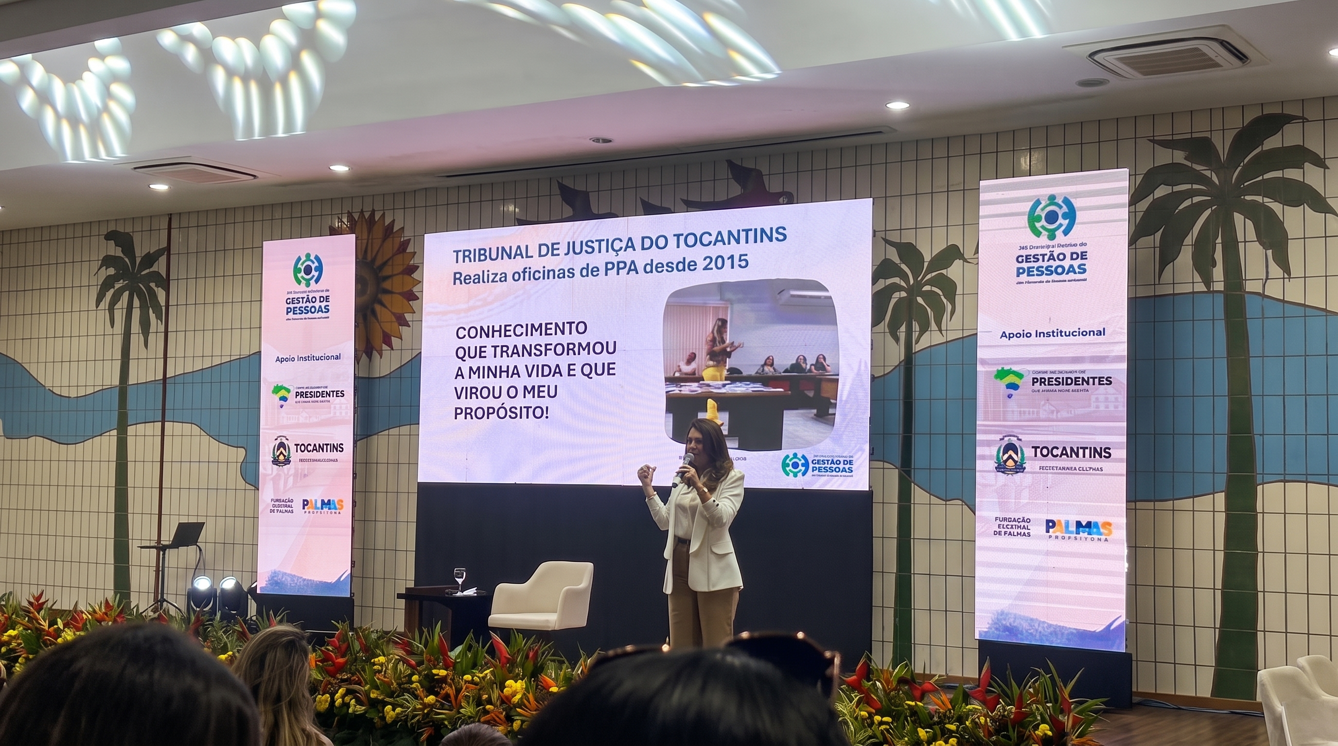 Fotografia da palestrante Cristina Prestes no XVI Encontro Nacional de Gestão de Pessoas dos Tribunais de Contas do Brasil. No painel ao fundo, lê-se o título "TRIBUNAL DE JUSTIÇA DO TOCANTINS Realiza oficinas de PPA desde 2015", acompanhado da frase "CONHECIMENTO QUE TRANSFORMOU A MINHA VIDA E QUE VIROU O MEU PROPÓSITO!"