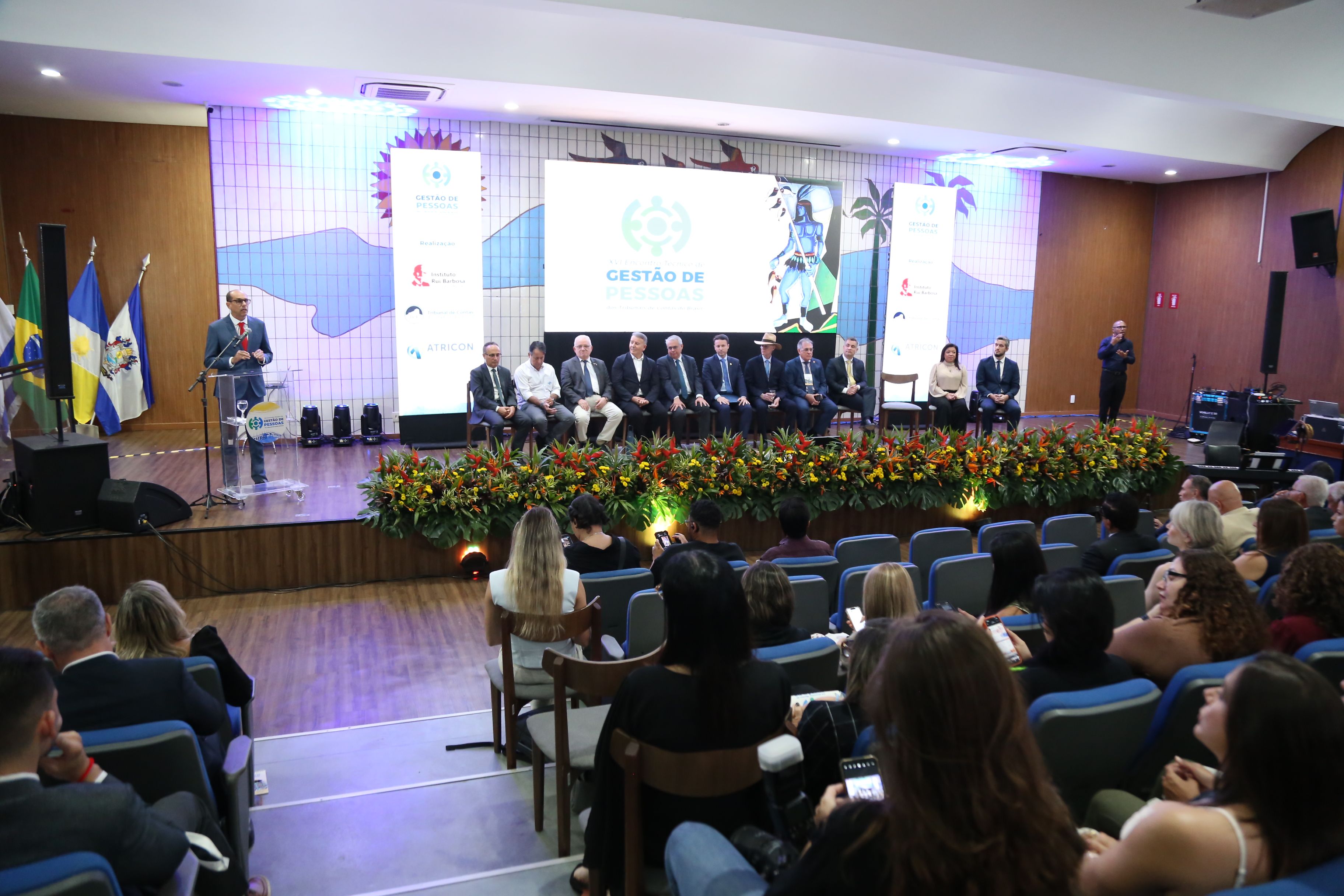 Fotografia da cerimônia de abertura do XVI Encontro Nacional de Gestão de Pessoas dos Tribunais de Contas do Brasil, realizado em Palmas. A imagem mostra o auditório lotado, visto da plateia para o palco. No palco, um homem fala ao microfone em um púlpito com o logotipo do evento. À direita, uma mesa de honra composta por várias autoridades.