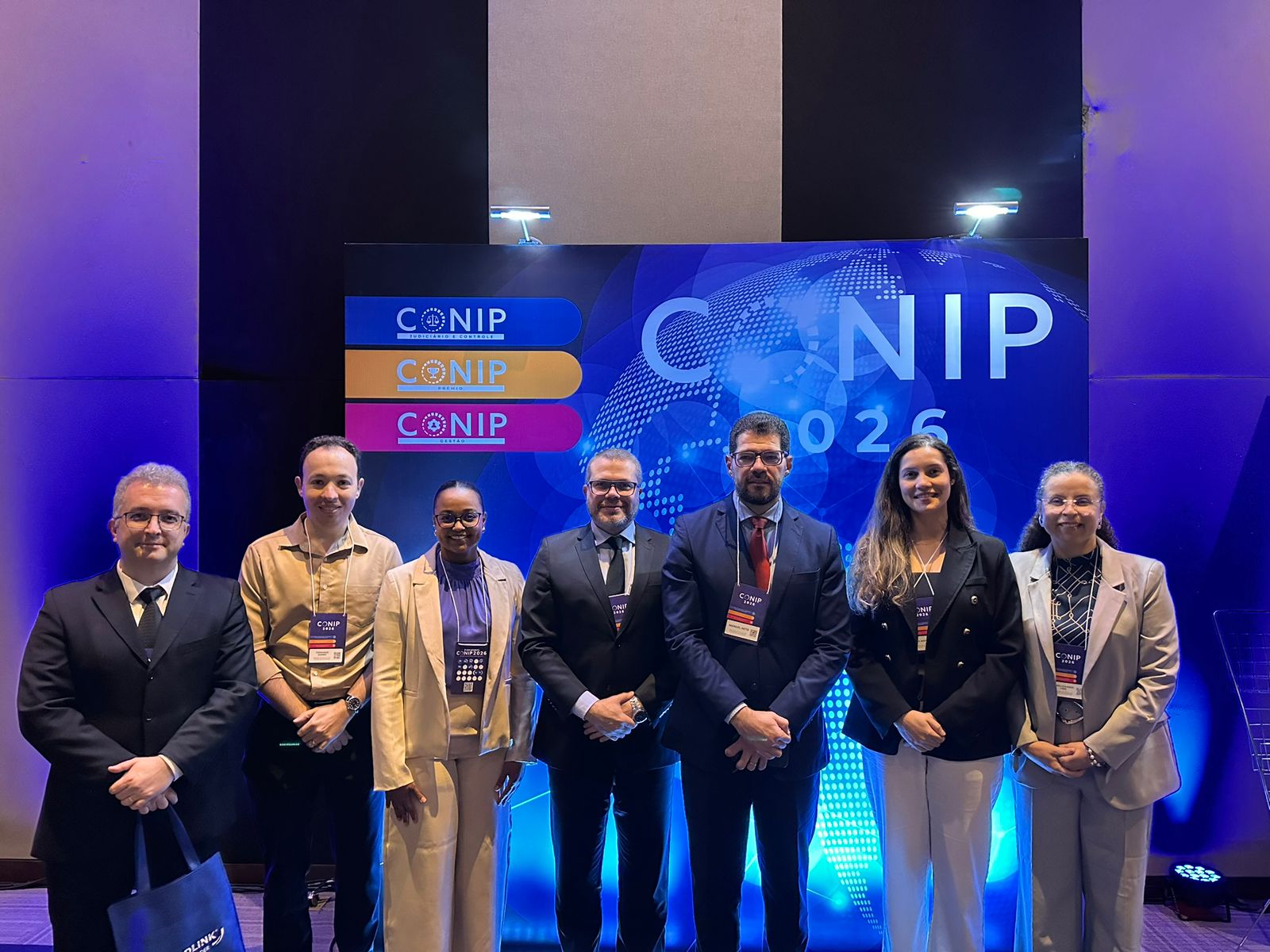 Foto mostra sete participantes do Conip Jud 2026 reunidos no palco, diante do painel do evento, em Brasília (DF). Entre eles, estão representantes da Corregedoria-Geral da Justiça do Tocantins (CGJUS), que participam do congresso realizado nos dias 2 e 3 de setembro.