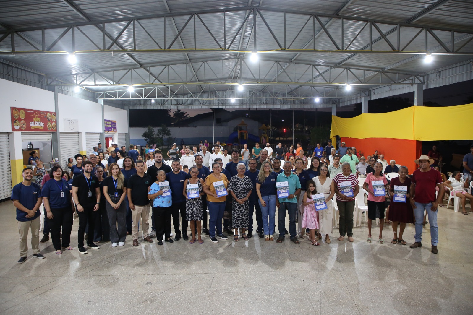A imagem mostra dezenas de pessoas reunidas em um espaço coberto durante a entrega de títulos de regularização fundiária. Na parte da frente, algumas pessoas seguram os documentos recebidos.