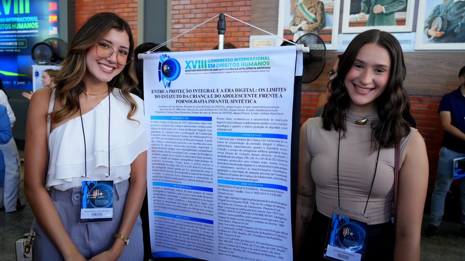 A imagem mostra duas mulheres posando para uma foto, uma de cada lado de um banner que apresenta informações sobre o XVIII Congresso Internacional em Direitos Humanos.