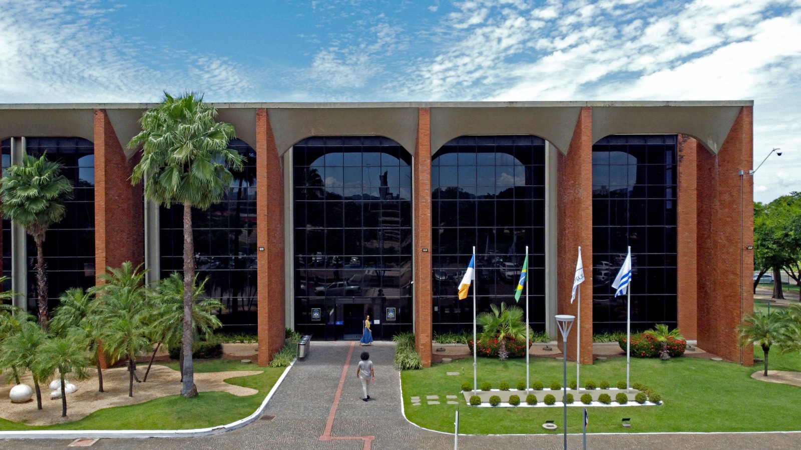 A imagem mostra a fachada do Tribunal de Justiça do Estado do Tocantins (Palácio Rio Tocantins), com arquitetura imponente em tijolos aparentes e grandes pilares verticais. O prédio é cercado por jardins, com vegetação ornamental.
