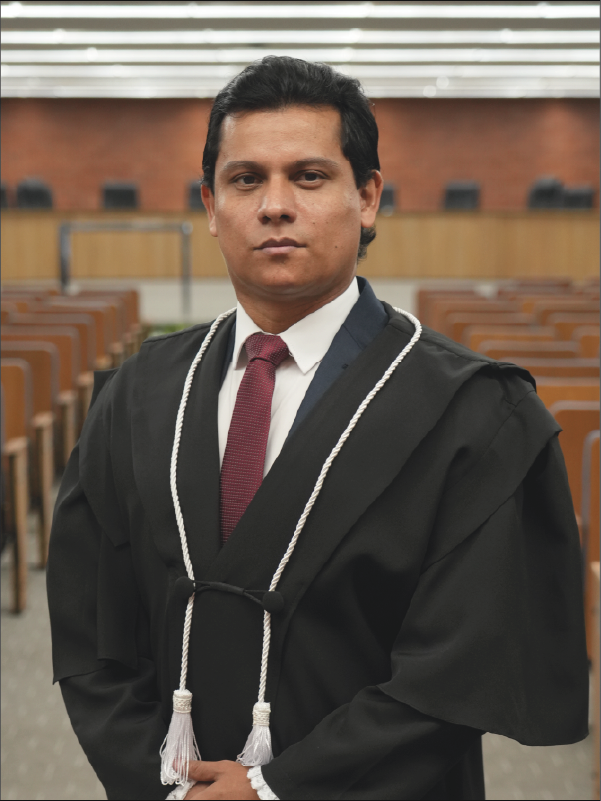 Homem em pé, em primeiro plano, olhando para a câmera, vestindo toga preta com cordões brancos, camisa branca e gravata em tom vermelho. Mantém as mãos unidas à frente do corpo. Ao fundo, aparece o plenário, com fileiras de cadeiras. A fotografia, em formato vertical, destaca o magistrado ao centro da imagem.