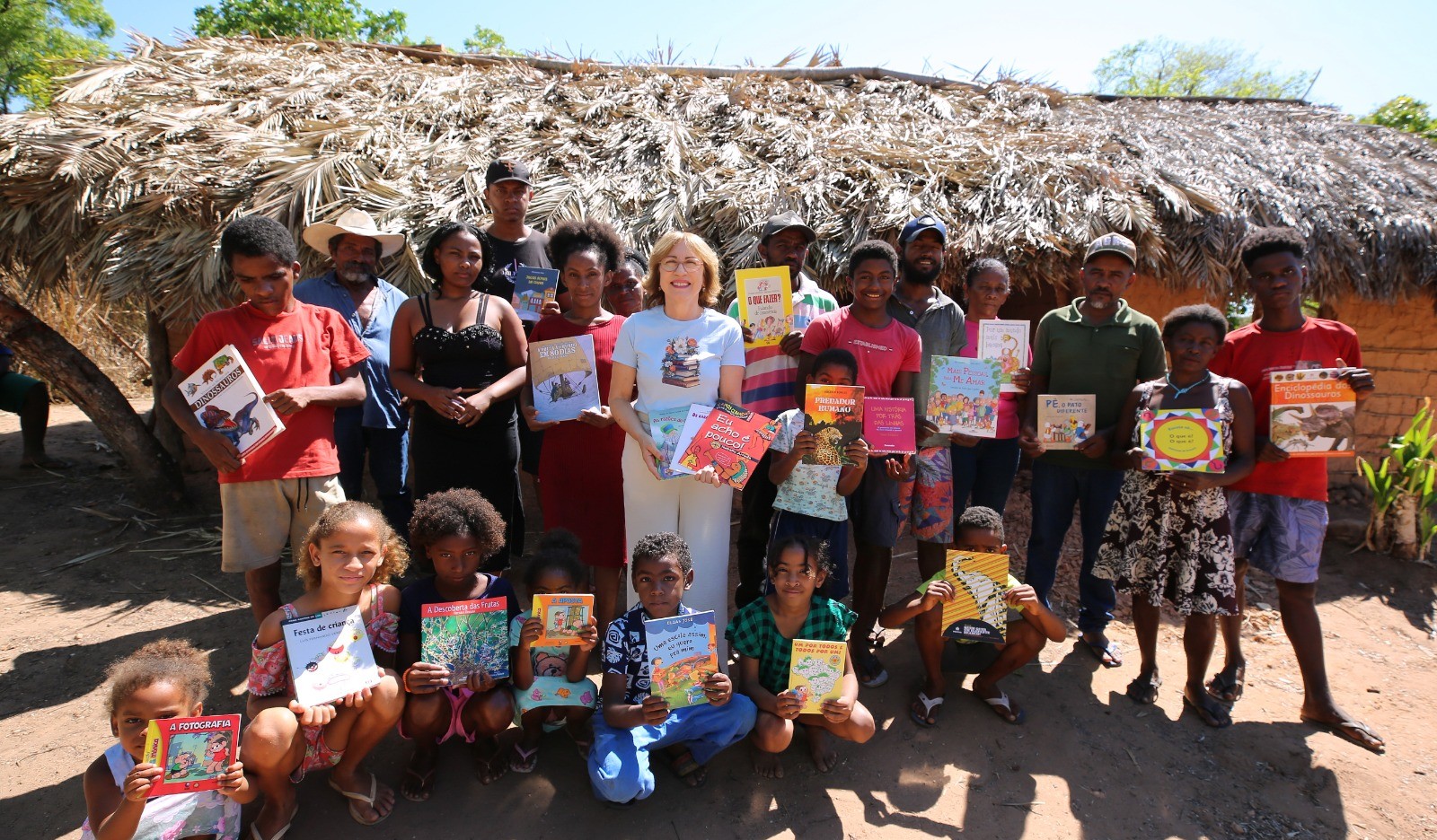 Fotografia mostra um grupo de adultos e crianças reunido em frente a uma casa de adobe coberta com palha, em uma comunidade rural. Várias pessoas seguram livros infantis e educativos. Ao centro, uma mulher de cabelos claros e óculos sorri enquanto segura diversos livros. Na parte da frente, crianças também exibem exemplares recebidos. A cena é ao ar livre, durante o dia, e registra um momento de entrega e incentivo à leitura.