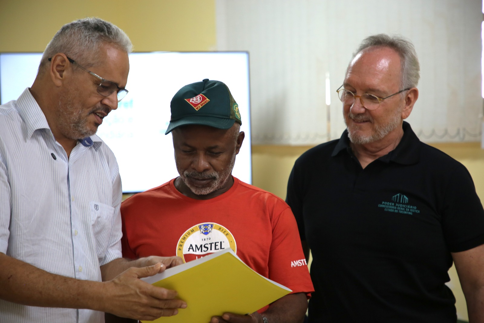 Fotografia mostra três homens em pé, em ambiente interno. À esquerda, um homem de camisa social clara e óculos segura uma pasta amarela aberta e mostra o documento aos demais. Ao centro, um homem de camiseta vermelha e boné verde observa atentamente o conteúdo. À direita, um homem de camisa polo preta, com a identificação do Poder Judiciário – Corregedoria-Geral da Justiça do Estado do Tocantins, acompanha o momento sorrindo. Ao fundo, há uma televisão e persianas claras.