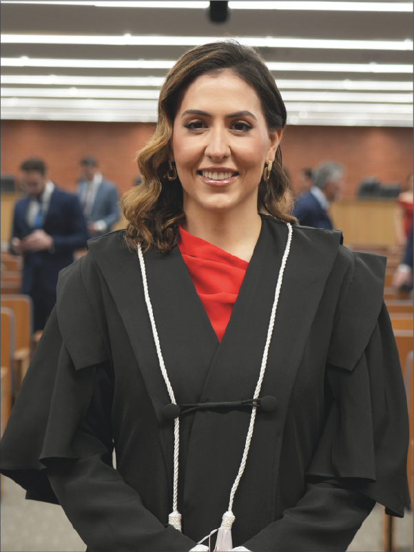 Mulher em pé, em primeiro plano, sorrindo para a câmera, vestindo toga preta com cordões brancos e blusa vermelha. Usa brincos dourados e mantém as mãos unidas à frente do corpo. Ao fundo, aparecem fileiras de cadeiras do plenário e algumas pessoas. A fotografia, em formato vertical, destaca a magistrada ao centro da imagem.