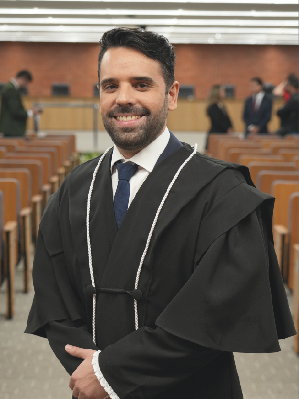 Homem em pé, em primeiro plano, sorrindo para a câmera, vestindo toga preta com cordões brancos, camisa branca e gravata azul-escura. Mantém as mãos unidas à frente do corpo. Ao fundo, aparecem fileiras de cadeiras do plenário e algumas pessoas. A fotografia, em formato vertical, destaca o magistrado ao centro da imagem.