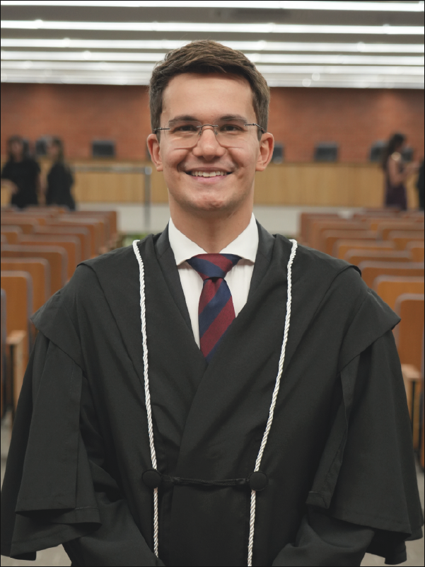 Homem em pé, em primeiro plano, sorrindo para a câmera, vestindo toga preta com cordões brancos, camisa branca, gravata listrada em azul e vermelho e óculos. Ao fundo, aparecem fileiras de cadeiras do plenário e algumas pessoas. A fotografia, em formato vertical, destaca o magistrado ao centro da imagem.