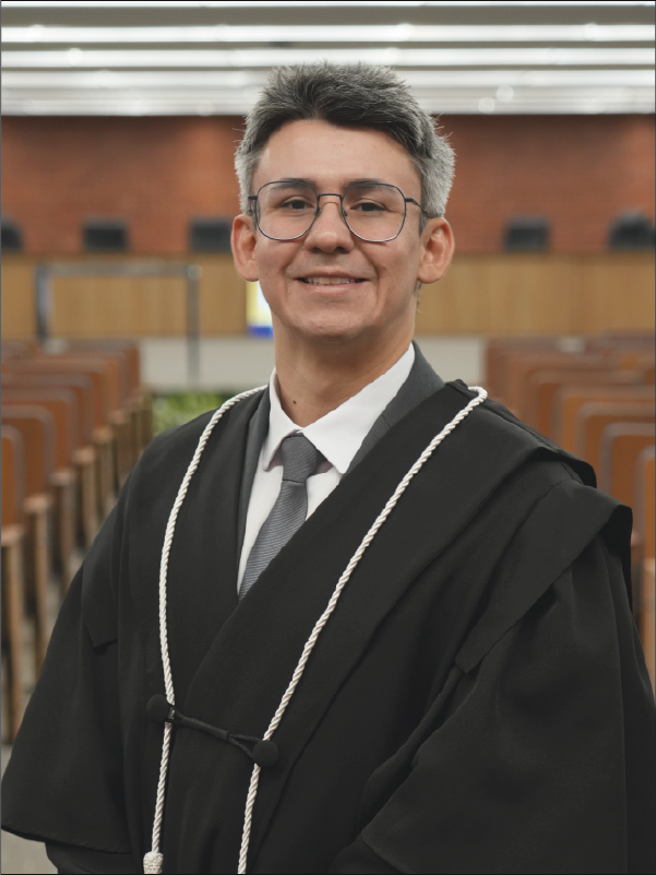 Homem em pé, em primeiro plano, sorrindo para a câmera, vestindo toga preta com cordões brancos, camisa branca, gravata cinza e óculos. Ao fundo, aparece o plenário, com fileiras de cadeiras. A fotografia, em formato vertical, destaca o magistrado ao centro da imagem.