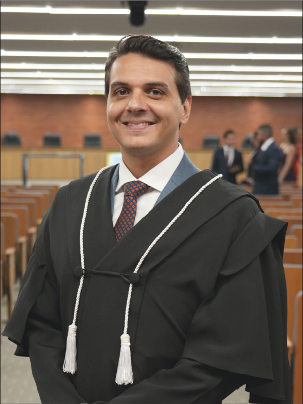 Homem em pé, em primeiro plano, sorrindo para a câmera, vestindo toga preta com cordões brancos, camisa branca e gravata estampada em tons de azul e laranja. Ao fundo, aparecem o plenário, com fileiras de cadeiras, e algumas pessoas. A fotografia, em formato vertical, destaca o magistrado ao centro da imagem.