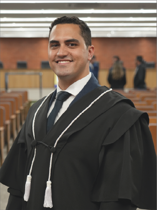 Homem em pé, em primeiro plano, sorrindo para a câmera, vestindo toga preta com cordões brancos, camisa branca e gravata escura. Ao fundo, aparecem o plenário, com fileiras de cadeiras, e algumas pessoas. A fotografia, em formato vertical, destaca o magistrado ao centro da imagem.