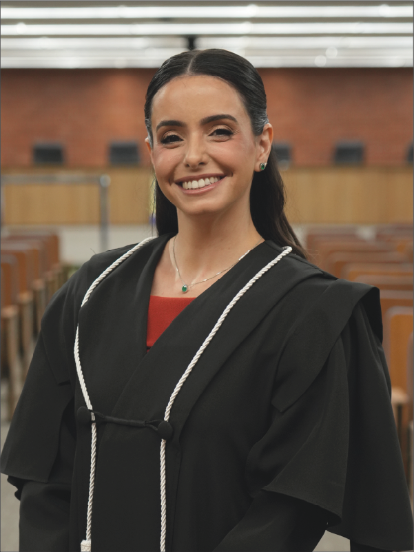 Mulher em pé, em primeiro plano, sorrindo para a câmera, vestindo toga preta com cordões brancos e blusa em tom vermelho. Usa brincos e colar com detalhes verdes. Ao fundo, aparece o plenário, com fileiras de cadeiras. A fotografia, em formato vertical, destaca a magistrada ao centro da imagem.