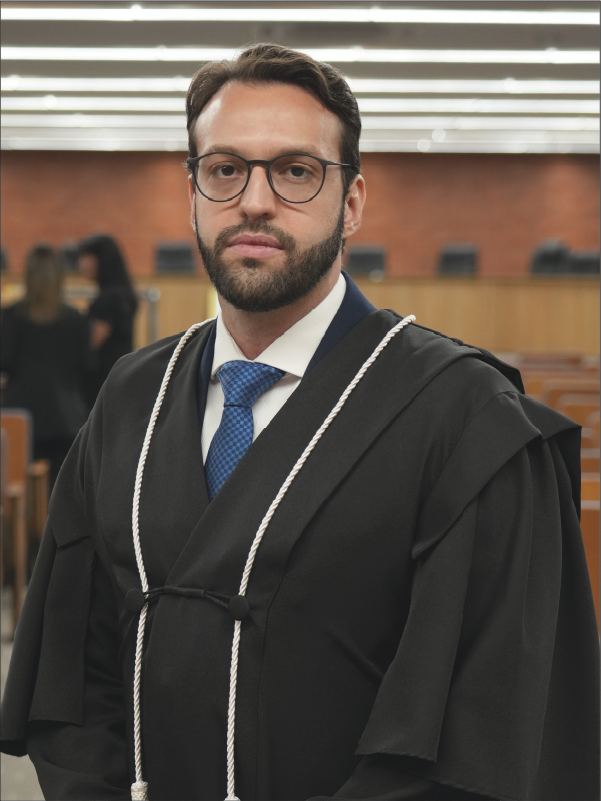 Homem em pé, em primeiro plano, olhando para a câmera, vestindo toga preta com cordões brancos, camisa branca, gravata azul e óculos de armação escura. Ao fundo, aparecem o plenário, com fileiras de cadeiras, e algumas pessoas. A fotografia, em formato vertical, destaca o magistrado ao centro da imagem.