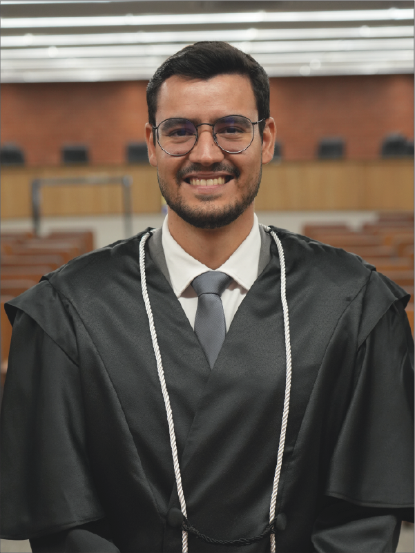 Homem em pé, em primeiro plano, sorrindo para a câmera, vestindo toga preta com cordões brancos, camisa branca, gravata cinza e óculos de armação escura. Ao fundo, aparece o plenário, com fileiras de cadeiras. A fotografia, em formato vertical, destaca o magistrado ao centro da imagem.