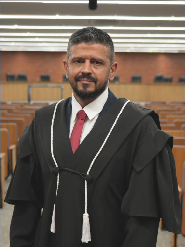 Homem em pé, em primeiro plano, olhando para a câmera, vestindo toga preta com cordões brancos, camisa branca e gravata vermelha. Ao fundo, aparece o plenário, com fileiras de cadeiras. A fotografia, em formato vertical, destaca o magistrado ao centro da imagem.