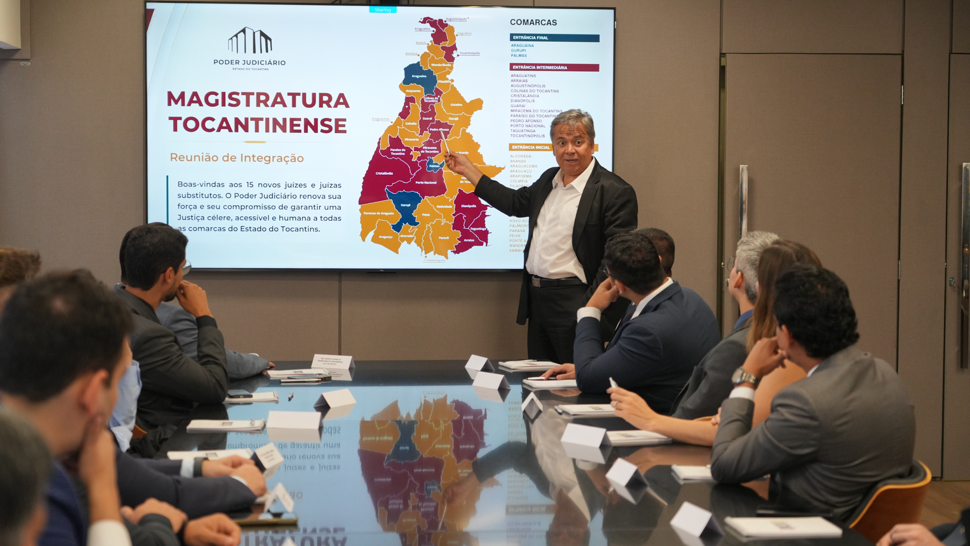 O juiz auxiliar da Presidência do TJTO, Arióstines Guimarães Vieira, apresenta aos novos magistrados e magistradas o mapa das comarcas tocantinenses durante reunião de integração. Ao fundo, a tela exibe a divisão das unidades judiciais por entrância.