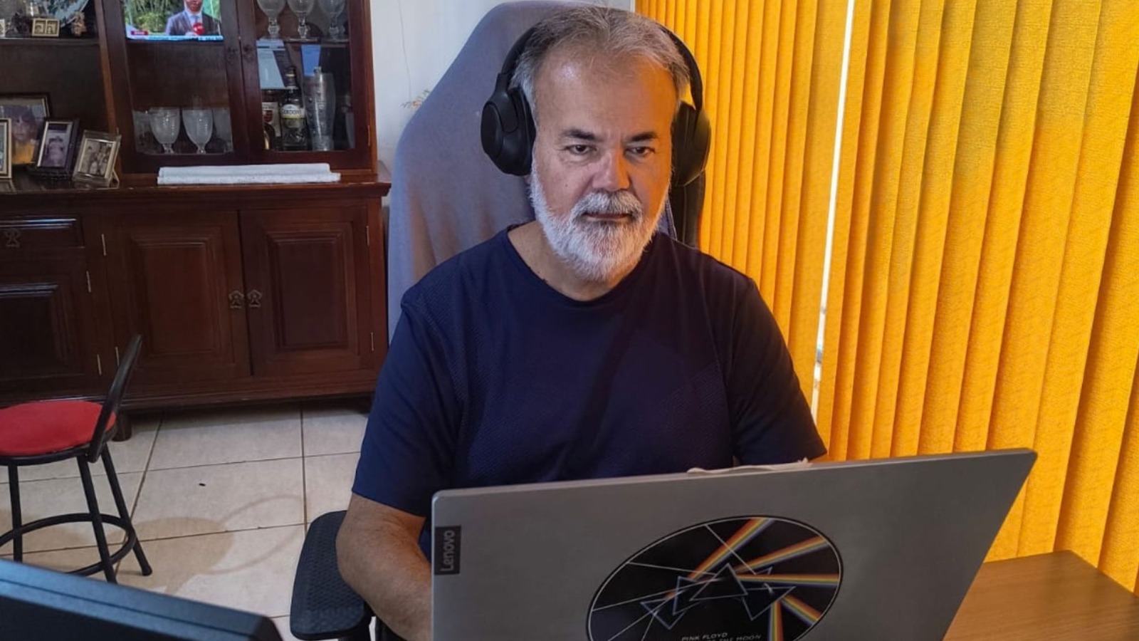 Fotografia doJosé Teixeira, homem de cabelo e barba grisalhos, usando fones de ouvido pretos e camiseta escura, sentado em frente a um notebook em teletrabalho.