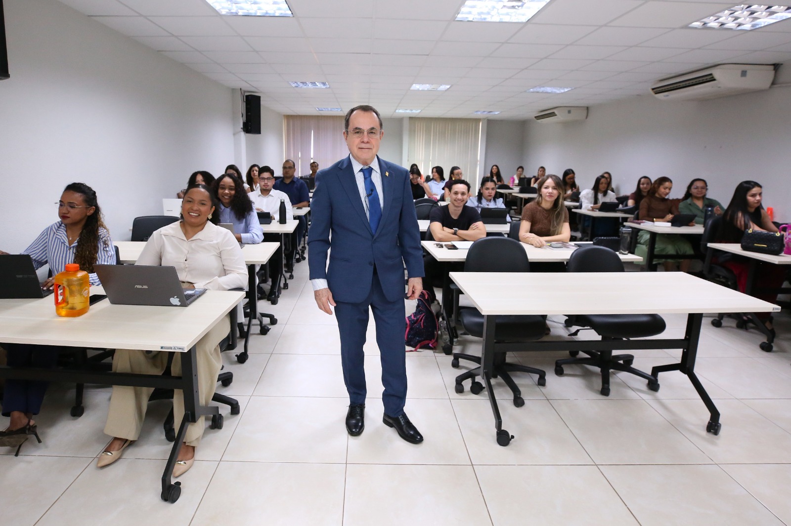 Fotografia: Desembargador Marco Villas Boas em pé no centro de sala de aula da Esmat, cercado por dezenas de estudantes residentes do PRJud sentados em bancadas com laptops abertos.