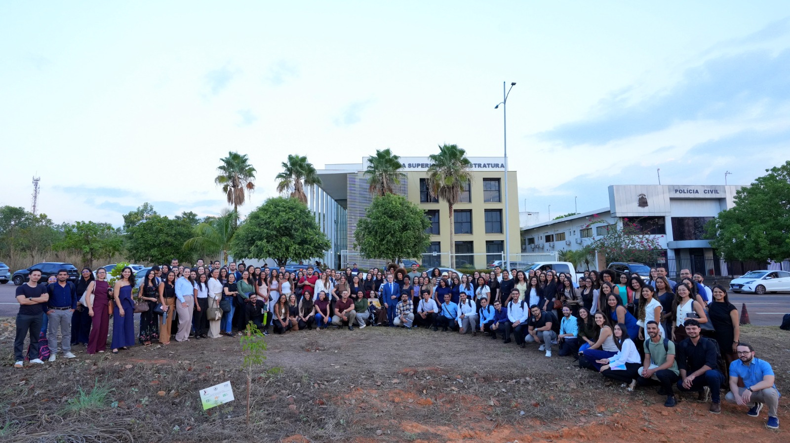 Fotografia de um grande grupo de residentes do PRJud reunidos ao ar livre em frente ao prédio da Esmat, posando juntos em área externa com vegetação e edifícios ao fundo.
