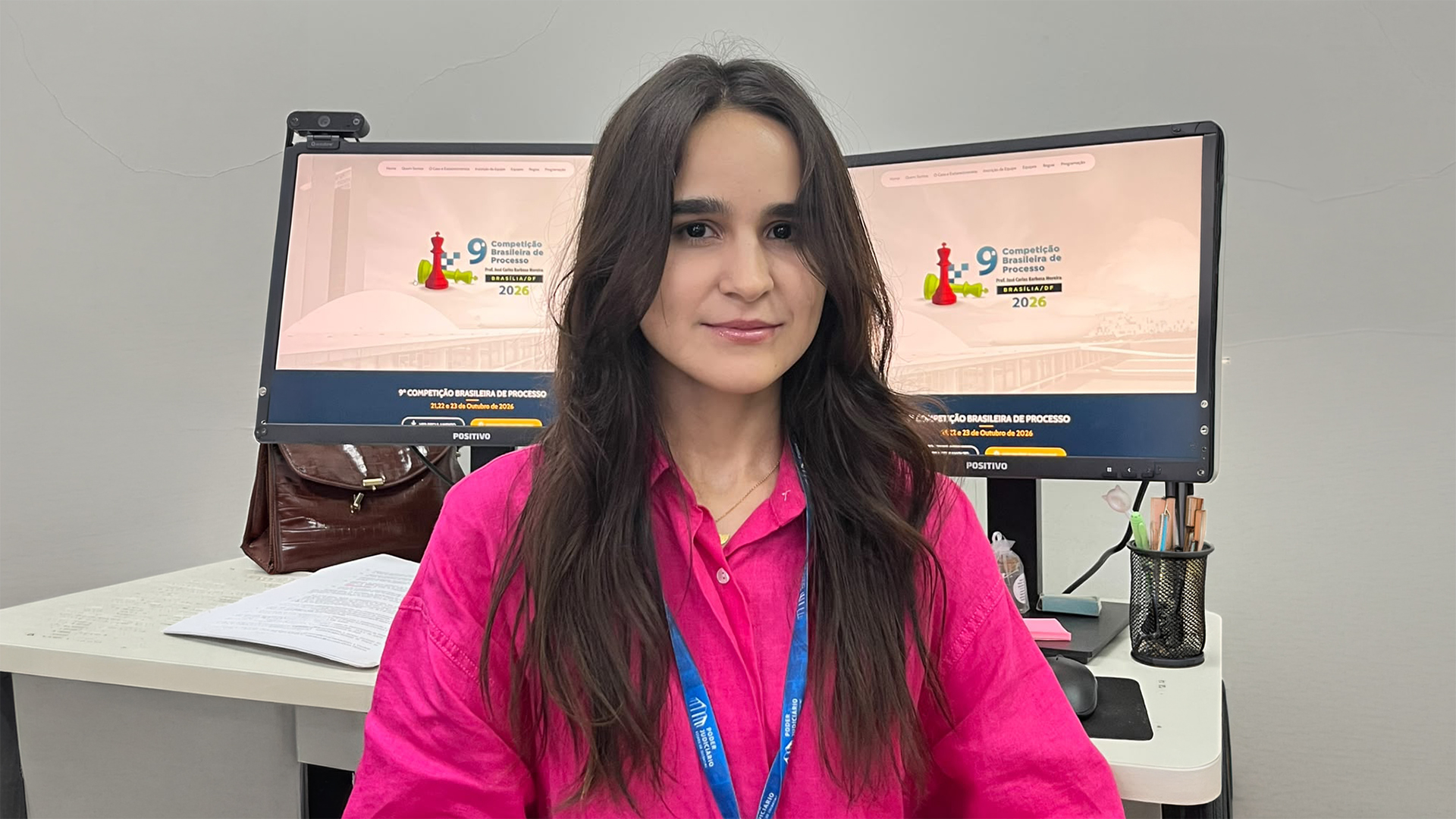 A imagem mostra uma mulher sentada em uma estação de trabalho, usando uma camisa rosa vibrante e um crachá azul do Poder Judiciário. Ao fundo, dois monitores exibem a identidade visual da 9ª Competição Brasileira de Processo, prevista para outubro de 2026, em Brasília/DF. Sobre um dos monitores há uma webcam, e a mesa possui documentos e materiais de escritório. O ambiente é interno, com paredes claras e iluminação suave.