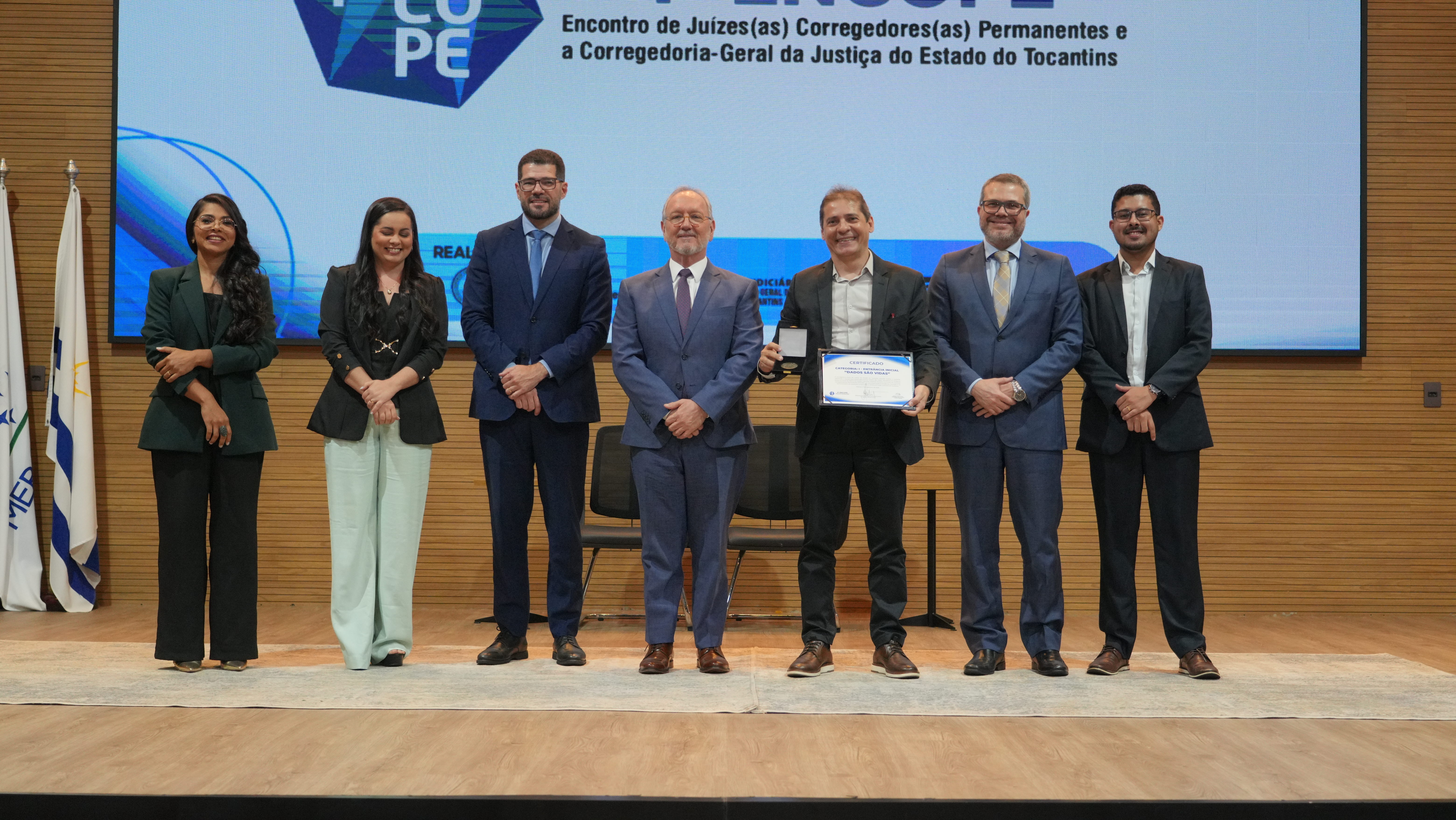 A imagem mostra sete pessoas reunidas no palco durante a entrega do 4º Prêmio Gestão Judiciária Ministro Sálvio de Figueiredo Teixeira; um dos homens segura o troféu e o certificado da premiação. Ao fundo, um telão exibe a identidade visual do IV Encontro de Juízas e Juízes Corregedores Permanentes (Encope).