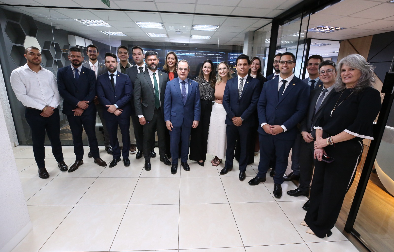 Grupo de pessoas, entre homens e mulheres, posa para foto em ambiente institucional, em frente a telas de computadores