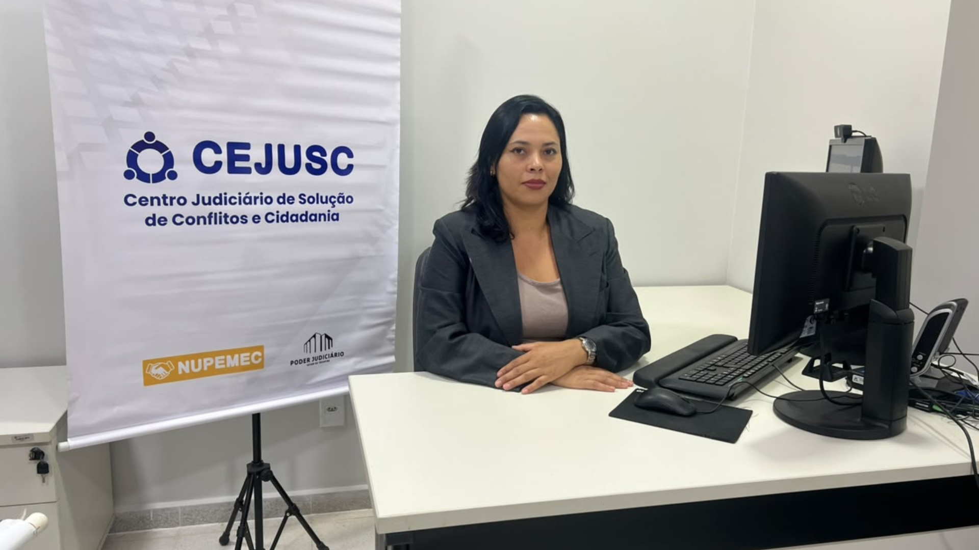 A imagem mostra Aline Lima de Jesus sentada à mesa, em frente a um computador, em um ambiente de trabalho do Cejusc. Ao lado, há um banner com a identificação do Centro Judiciário de Solução de Conflitos e Cidadania (Cejusc) e os logotipos do Nupemec e do Poder Judiciário.