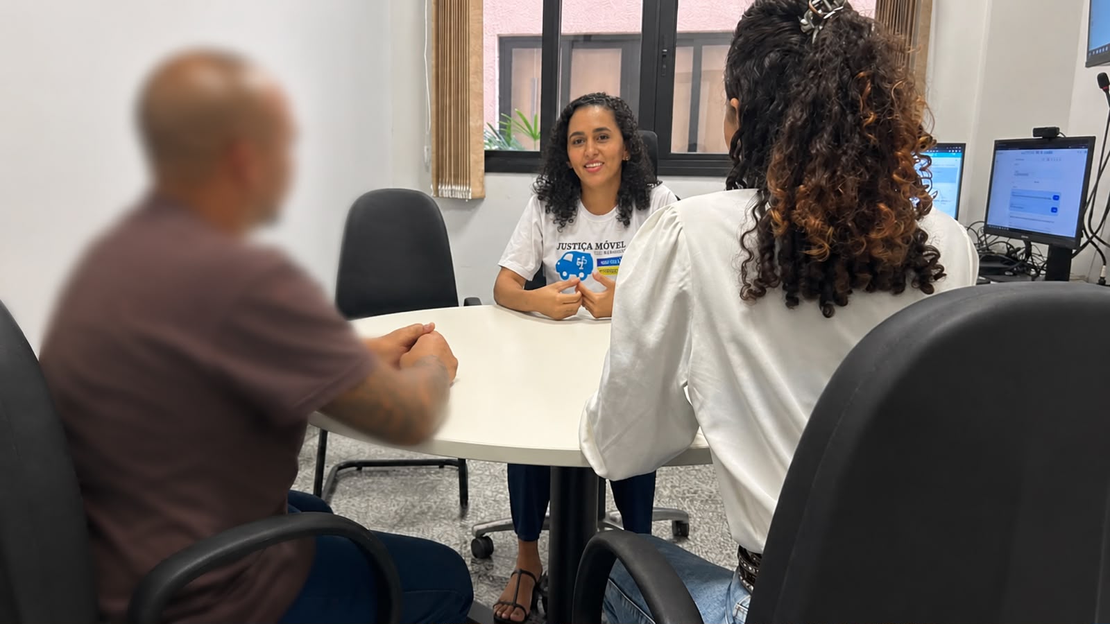 A imagem mostra Maria Luiza sentada à mesa, durante uma sessão de conciliação. Ela usa uma camiseta com a identificação da Justiça Móvel, enquanto duas pessoas aparecem de costas no primeiro plano.