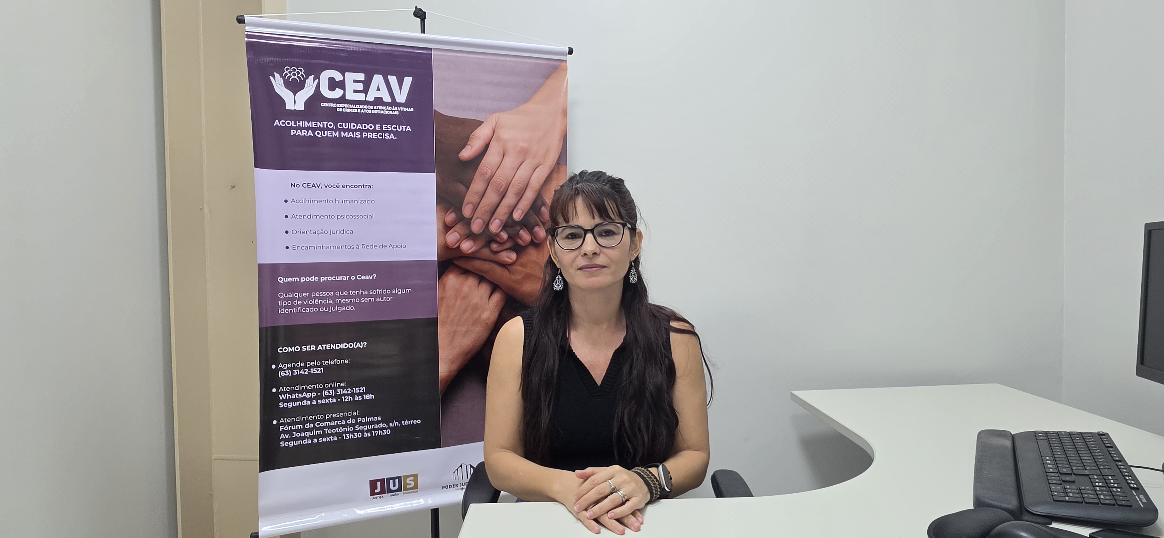 A imagem mostra uma mulher sentada atrás de uma mesa branca em um ambiente de atendimento, olhando para a câmera com as mãos apoiadas uma sobre a outra. Ao lado esquerdo, há um banner do CEAV – Centro Especializado de Atenção às Vítimas de Crimes e Atos Infracionais, nas cores roxo e branco, com a frase “Acolhimento, cuidado e escuta para quem mais precisa”, além de informações sobre os serviços oferecidos e formas de atendimento.  