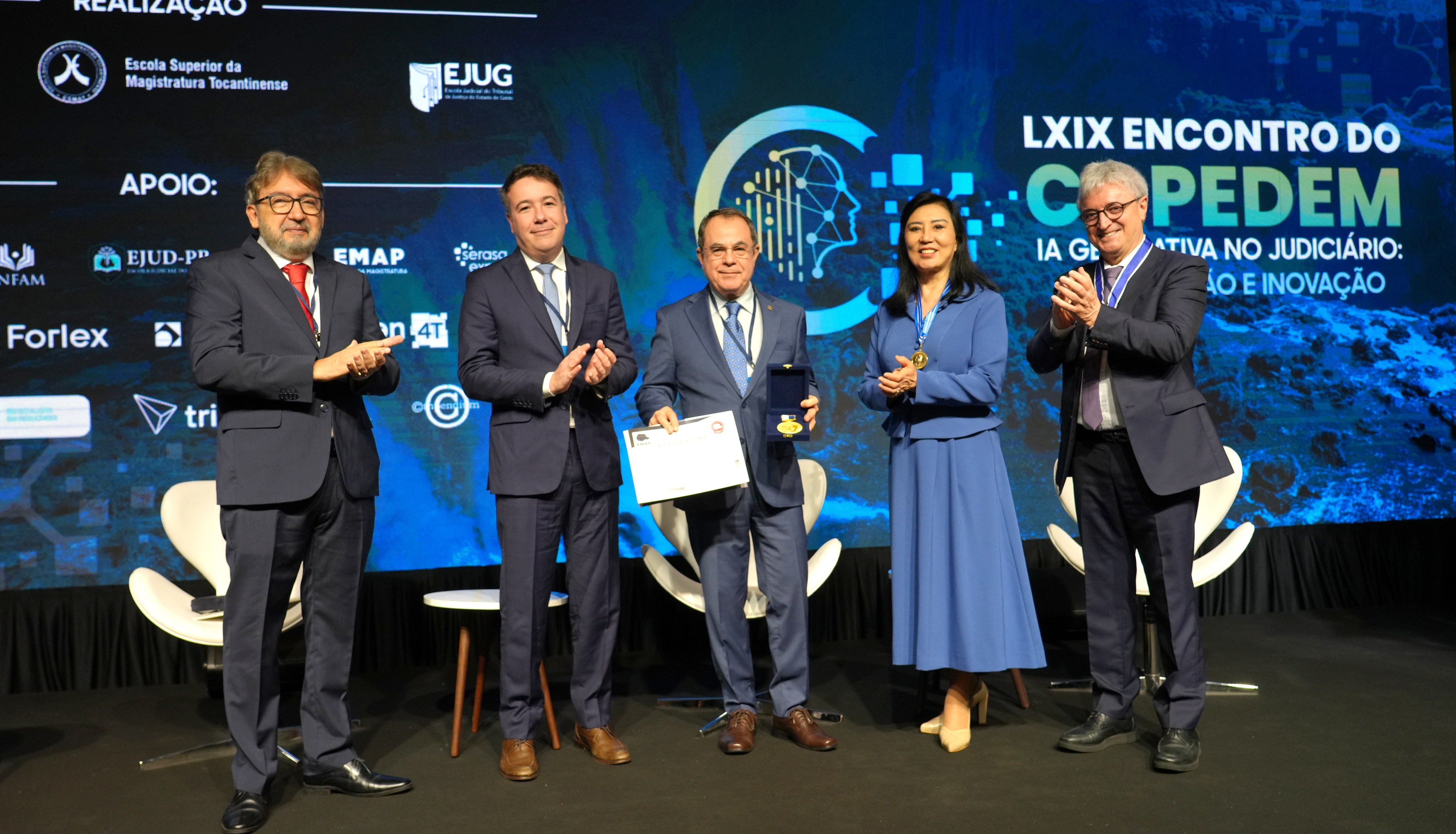 A imagem mostra cinco pessoas em um palco, durante o LXIX Encontro do Copedem, com o desembargador Marco Villas Boas ao centro segurando um certificado e uma medalha recebidos em homenagem.