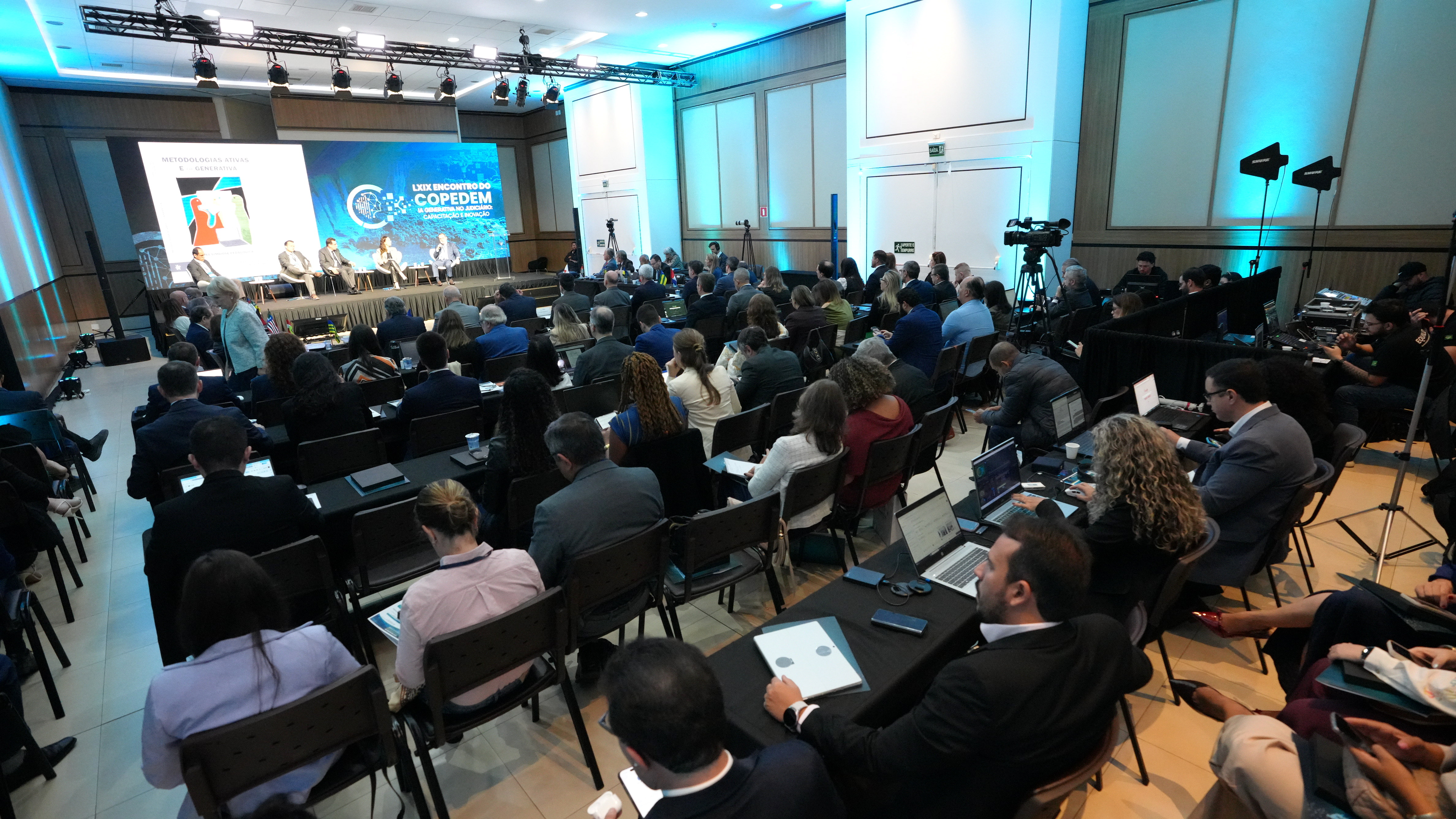 A imagem mostra participantes do LXIX Encontro do Copedem sentados em um auditório, acompanhando uma apresentação sobre Inteligência Artificial Generativa no Judiciário. Ao fundo, um painel exibe a identificação do evento e, no palco, palestrantes participam de um painel diante do público.