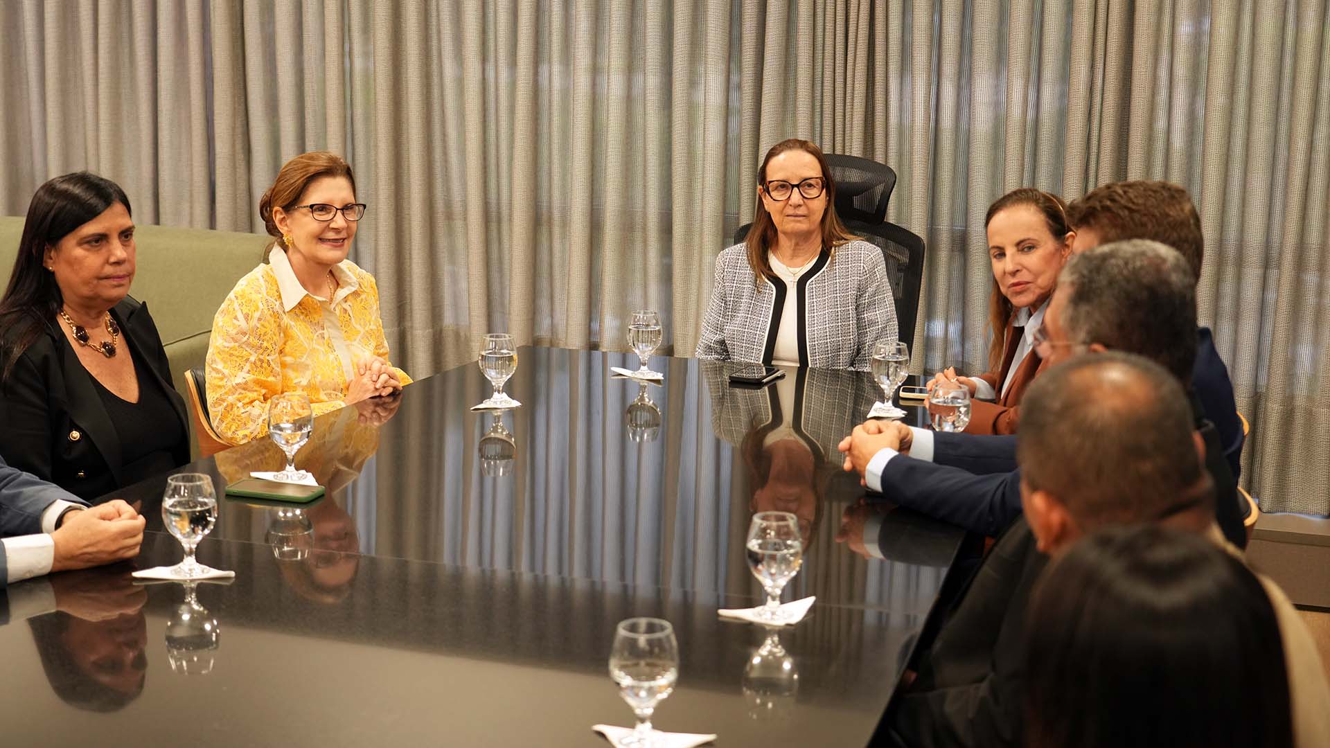 A imagem mostra uma reunião institucional em uma sala de reuniões do Tribunal de Justiça do Tocantins. Ao redor de uma mesa retangular de vidro escuro, a presidente do TJTO, desembargadora Maysa Vendramini Rosal, recebe a presidente do Superior Tribunal Militar, ministra Maria Elizabeth Guimarães Teixeira, acompanhadas de magistrados e magistradas. Sobre a mesa há taças com água e celulares. Ao fundo, cortinas em tons neutros cobrem toda a parede, compondo um ambiente sóbrio e formal.