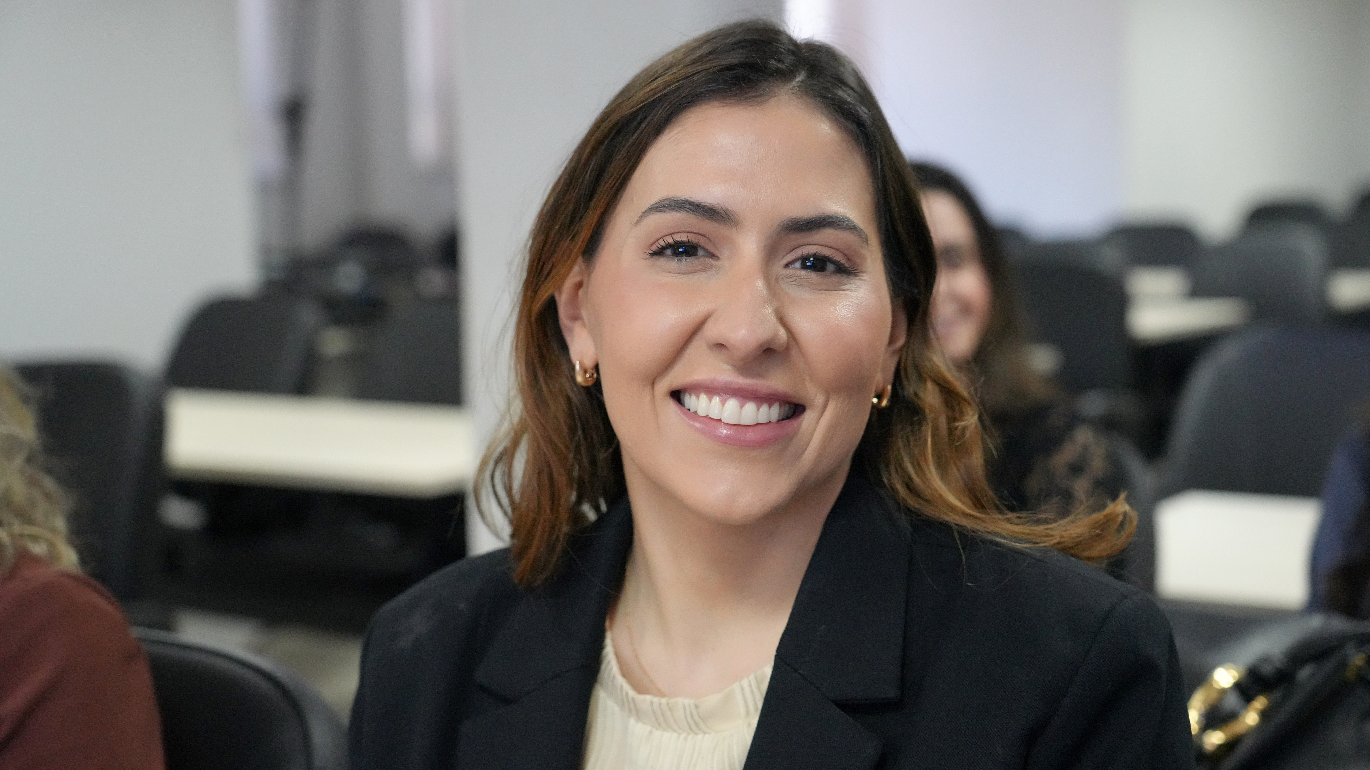 A juíza Ivie sorri para a câmera em um ambiente com outras pessoas e cadeiras ao fundo. Ela veste blazer preto sobre uma blusa clara.