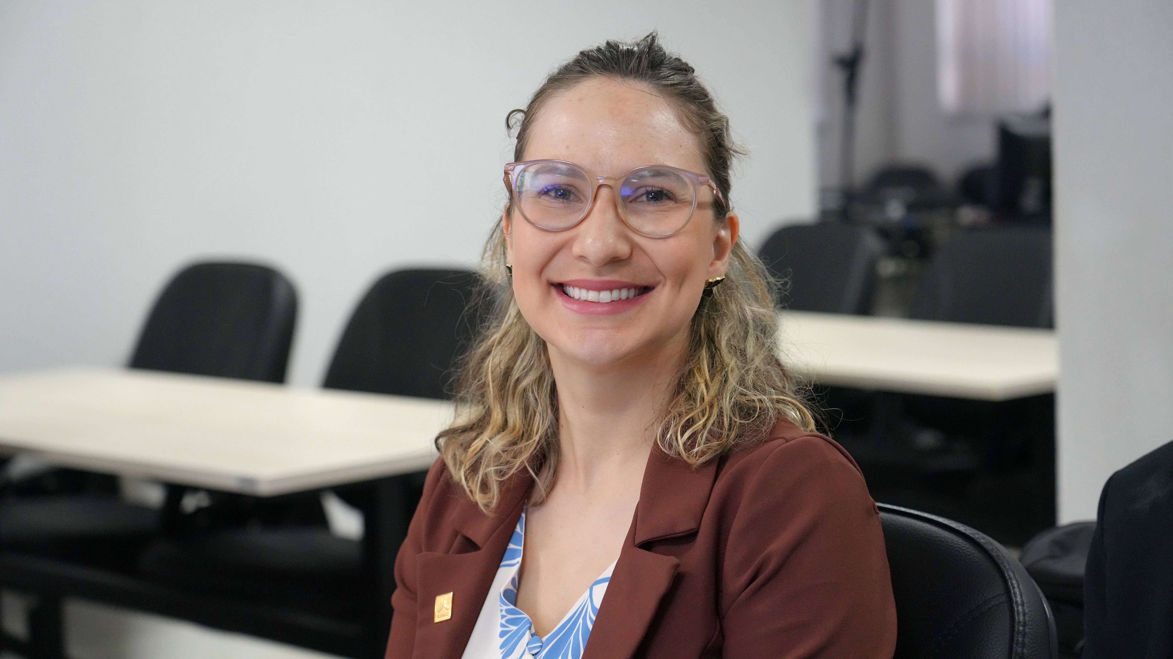 A juíza Gabriela aparece sentada e sorrindo para a câmera. Ela usa óculos de armação clara e blazer marrom sobre uma blusa estampada em azul e branco. Mesas e cadeiras aparecem ao fundo.