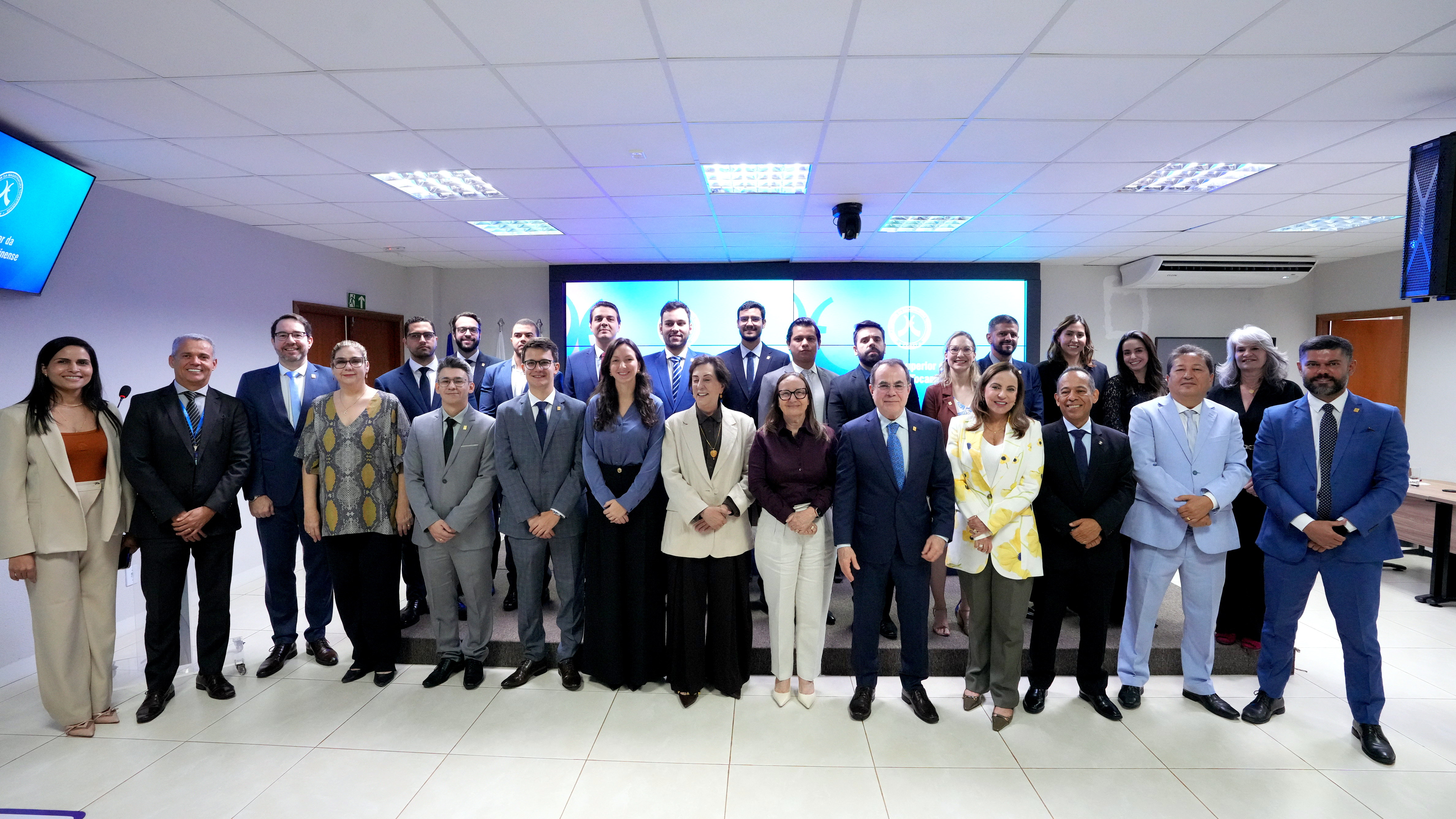 Magistrados e magistradas posam para foto em duas fileiras, em uma sala de eventos. A maioria veste trajes sociais. Ao fundo, há um painel de telas com elementos gráficos em tons de azul.