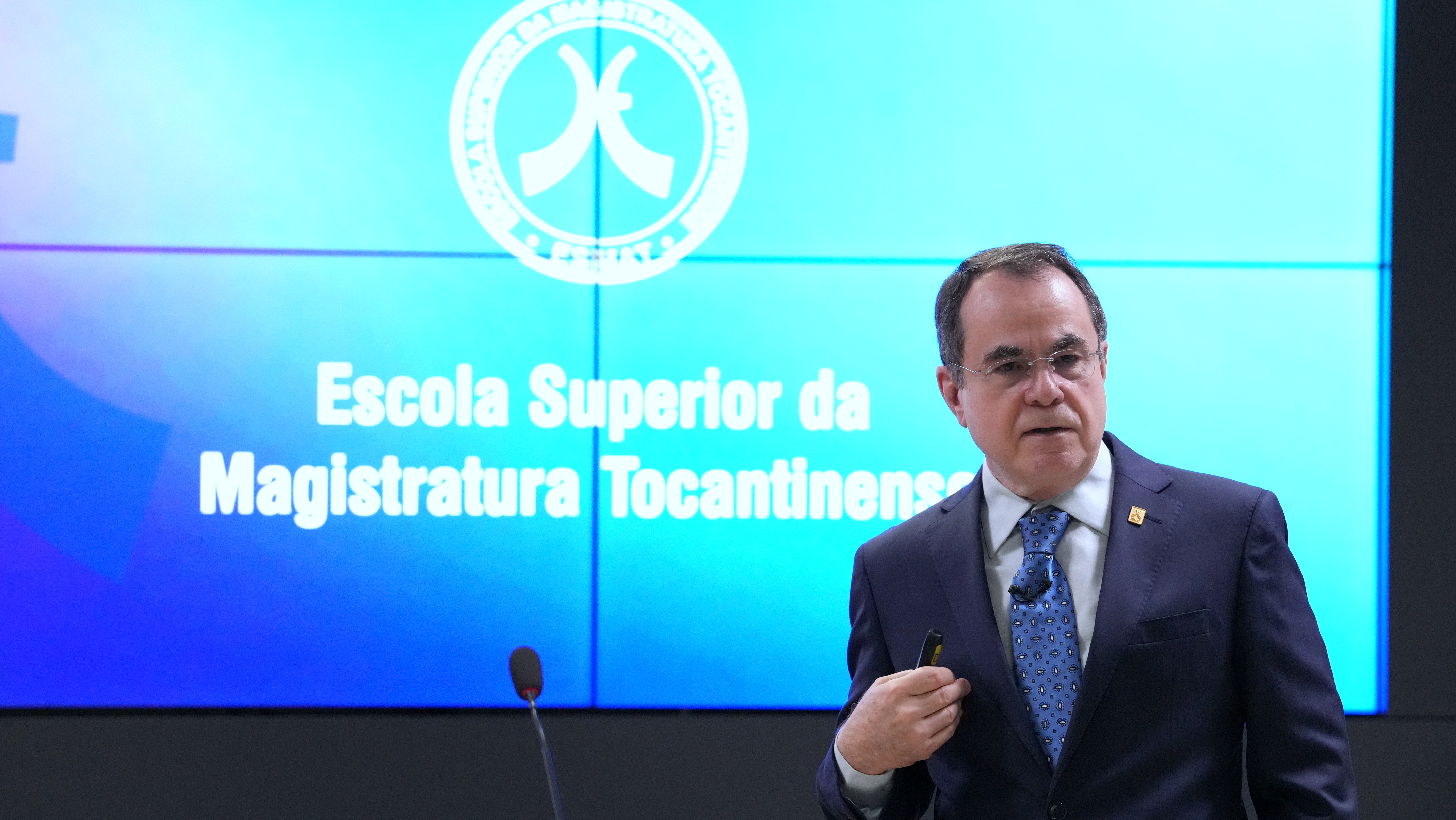 Desembargador Villas Boas aparece de frente, em primeiro plano, e ao fundo a logo da Esmat num telão em tons de azul
